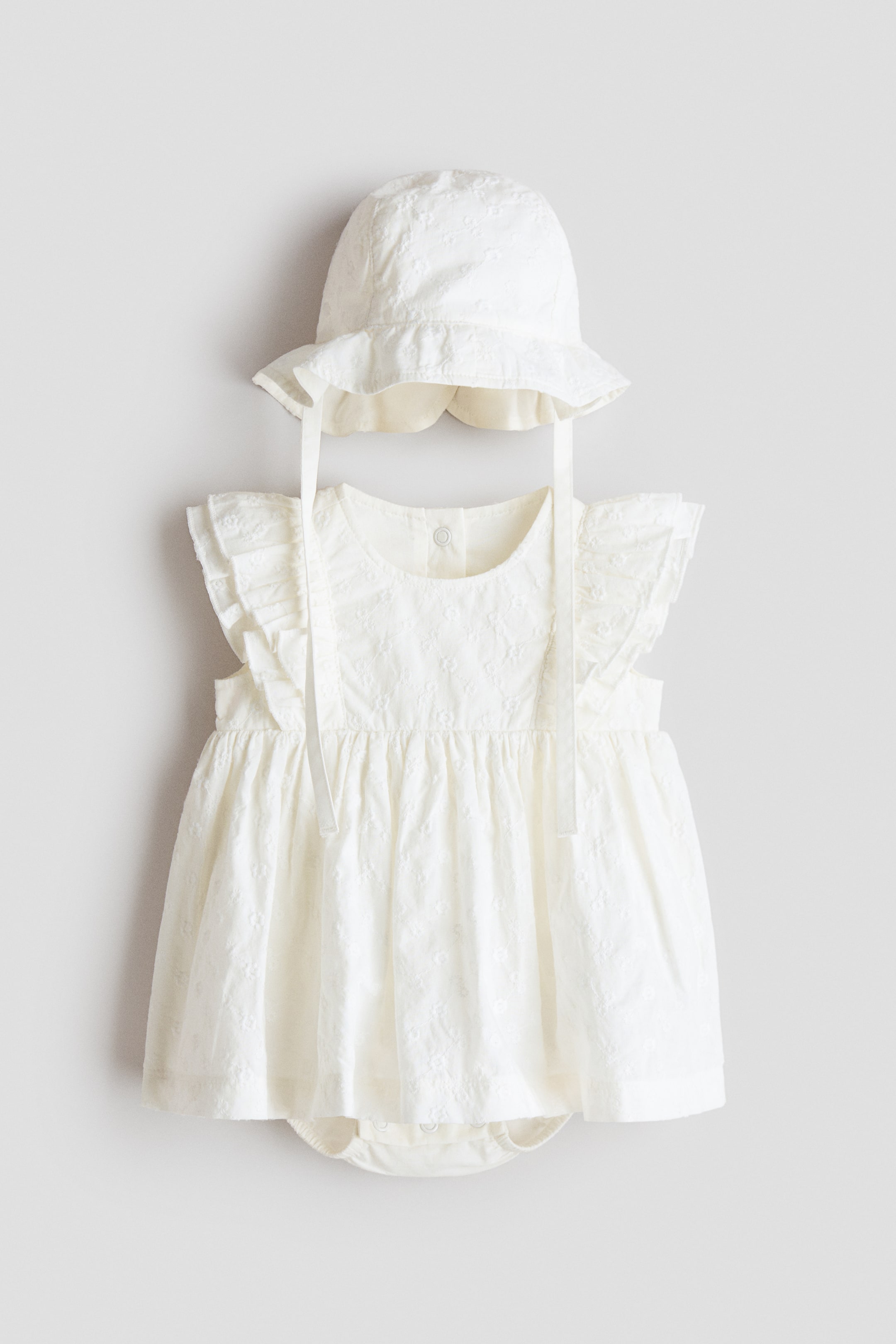 View larger image: An off-white cotton muslin baby's bodysuit dress and matching sun hat are presented, front view. The sleeveless dress features a round neckline, decorative frills over the shoulders, an all-over textured pattern and a gathered waistline with a flared skirt, revealing an integrated bodysuit beneath. The coordinating sun hat showcases a wavy brim and chin ties.