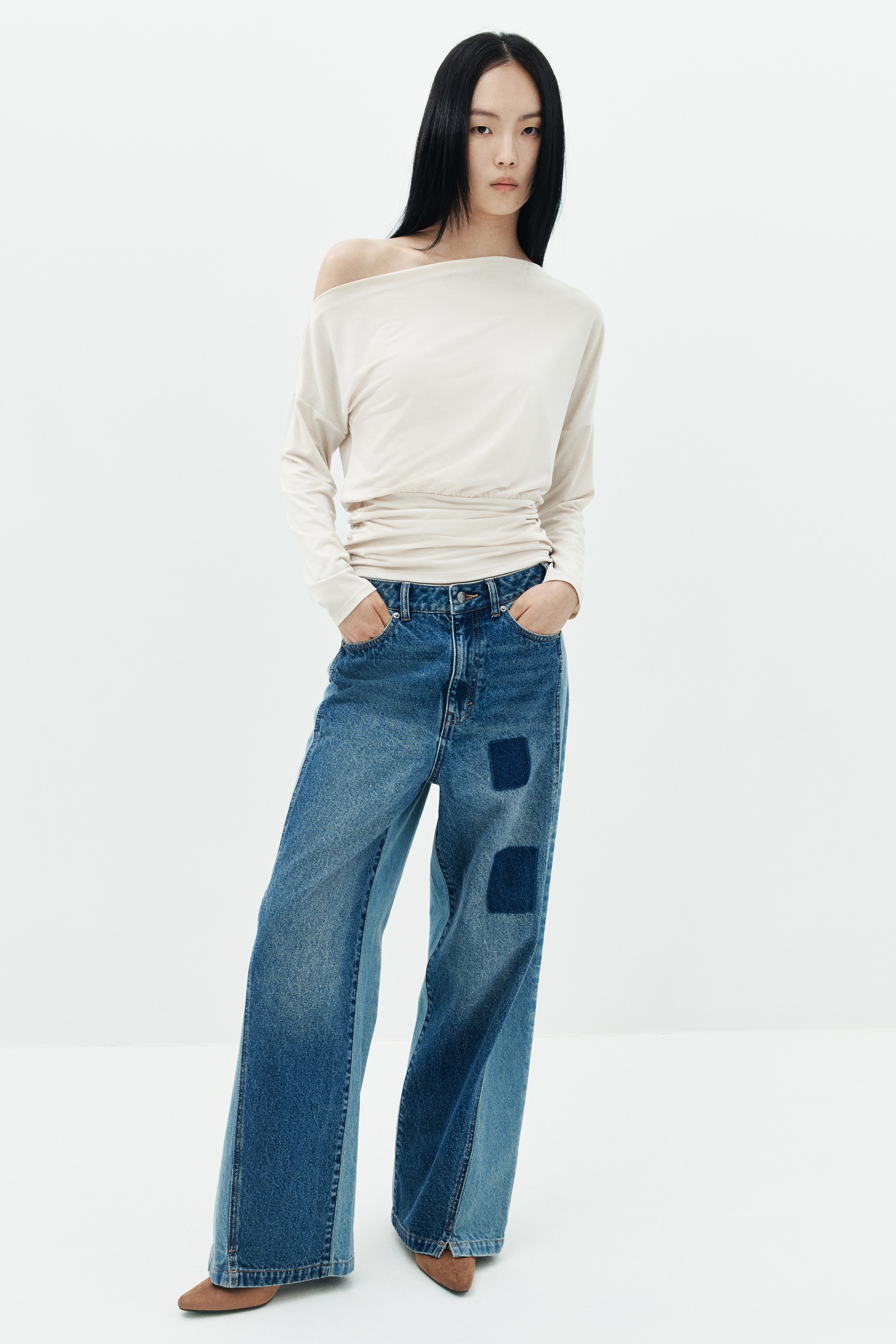 A pair of wide-leg jeans is facing front, showcasing a two-tone blue denim design with lighter-wash side panels and a waistband that contrasts with medium-wash front panels. The jeans feature two dark blue square patches on the left thigh and classic five-pocket styling.