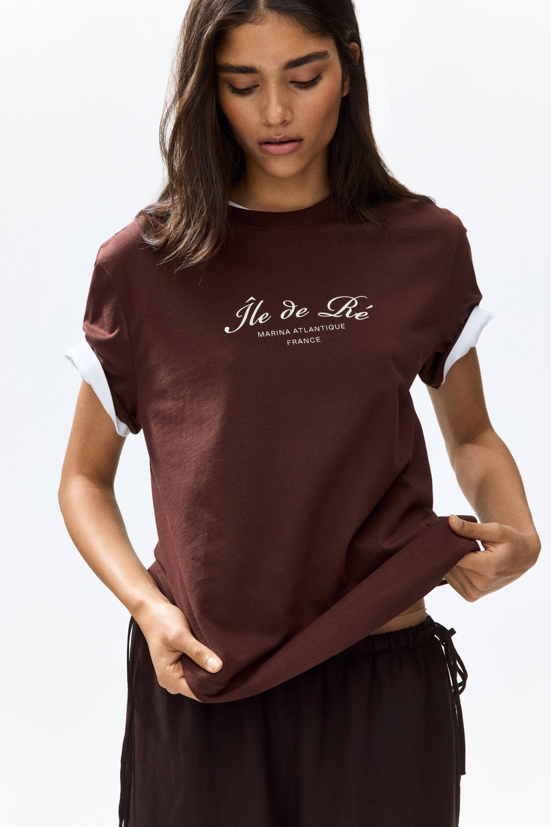 View larger image: The model is wearing a dark brown, soft cotton jersey T-shirt featuring a white "Île de Ré MARINA ATLANTIQUE FRANCE" print on the front. It comes with a round, rib-trimmed neckline and rolled short sleeves that reveal a white underlayer. The model is gently pulling the T-shirt's bottom hem with both hands.