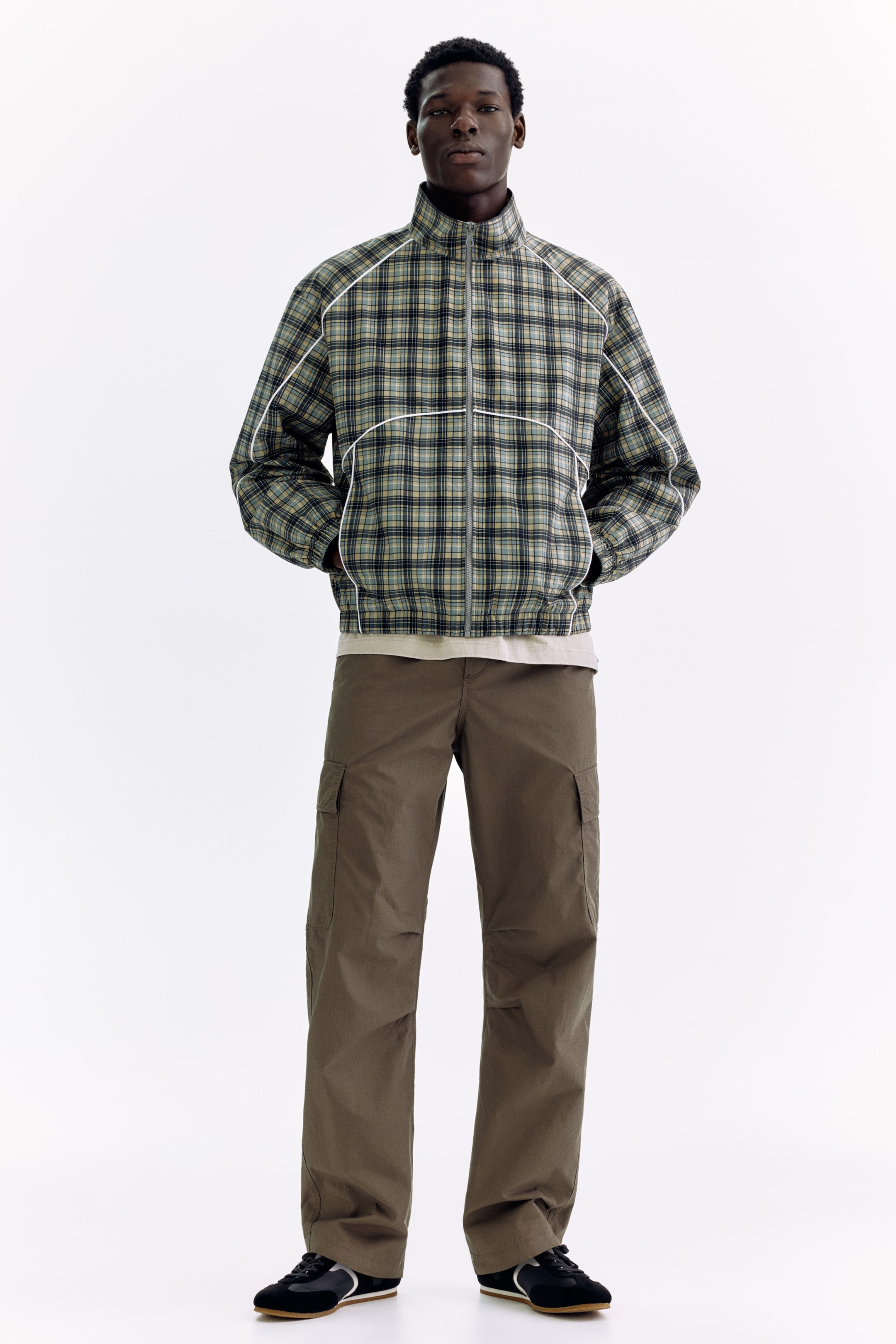 Agrandir l'image: Une veste à carreaux verte, noire et beige à fermeture éclair intégrale, avec un col montant et des détails de passepoil blanc sur les coutures raglan et la poitrine, est présentée de face. Un t-shirt beige clair est visible en superposition sous la veste, accompagné d'un pantalon cargo marron avec des poches latérales et des baskets noires avec des rayures blanches.