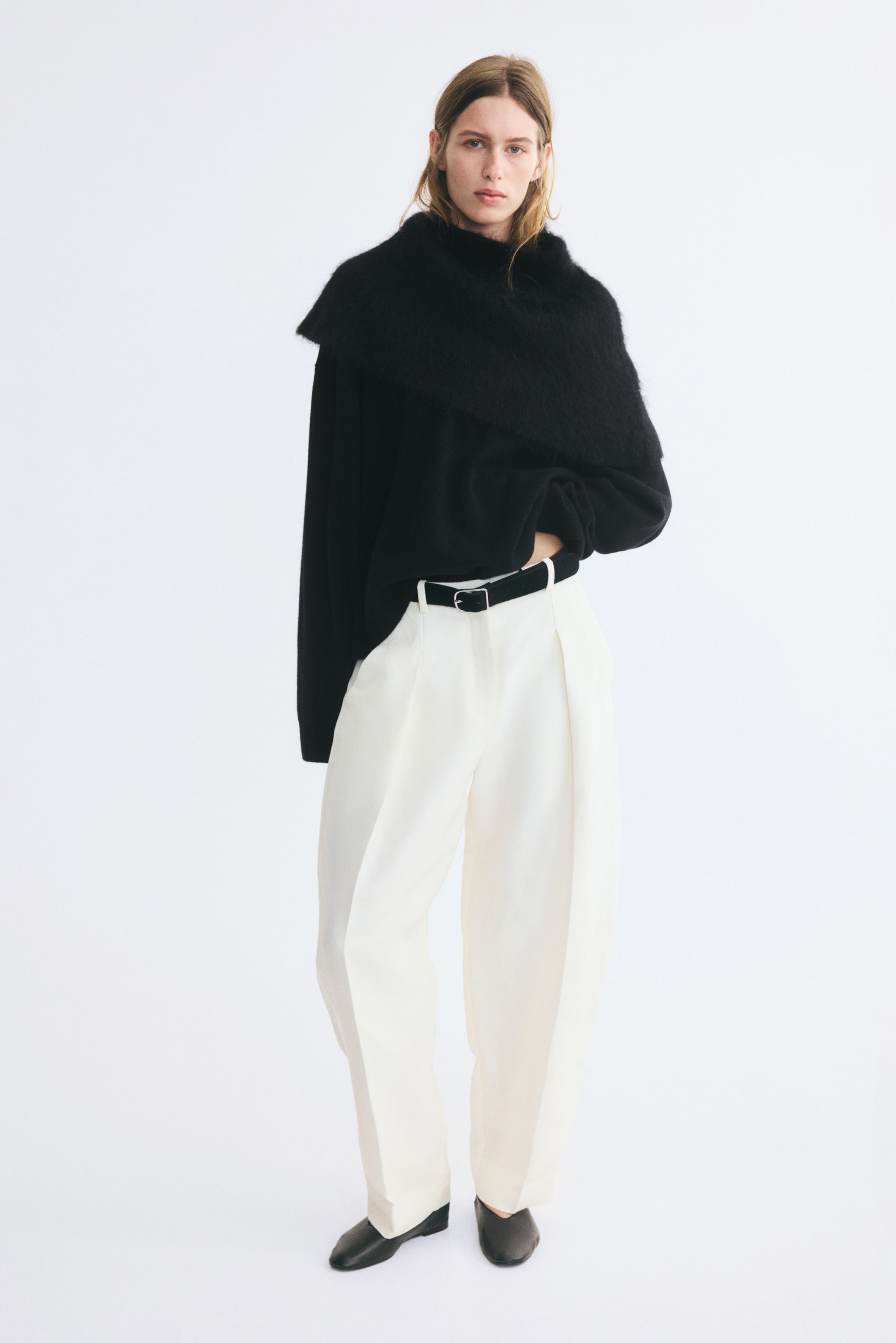 Visualizza l'immagine ingrandita: Un maglione nero oversize in maglia, con maniche lunghe, spalle scese e un colletto sovrapposto drappeggiato e morbido, è indossato dalla modella. Il maglione è leggermente infilato in vita in pantaloni bianchi plissettati a vita alta, abbinati a una cintura nera.
