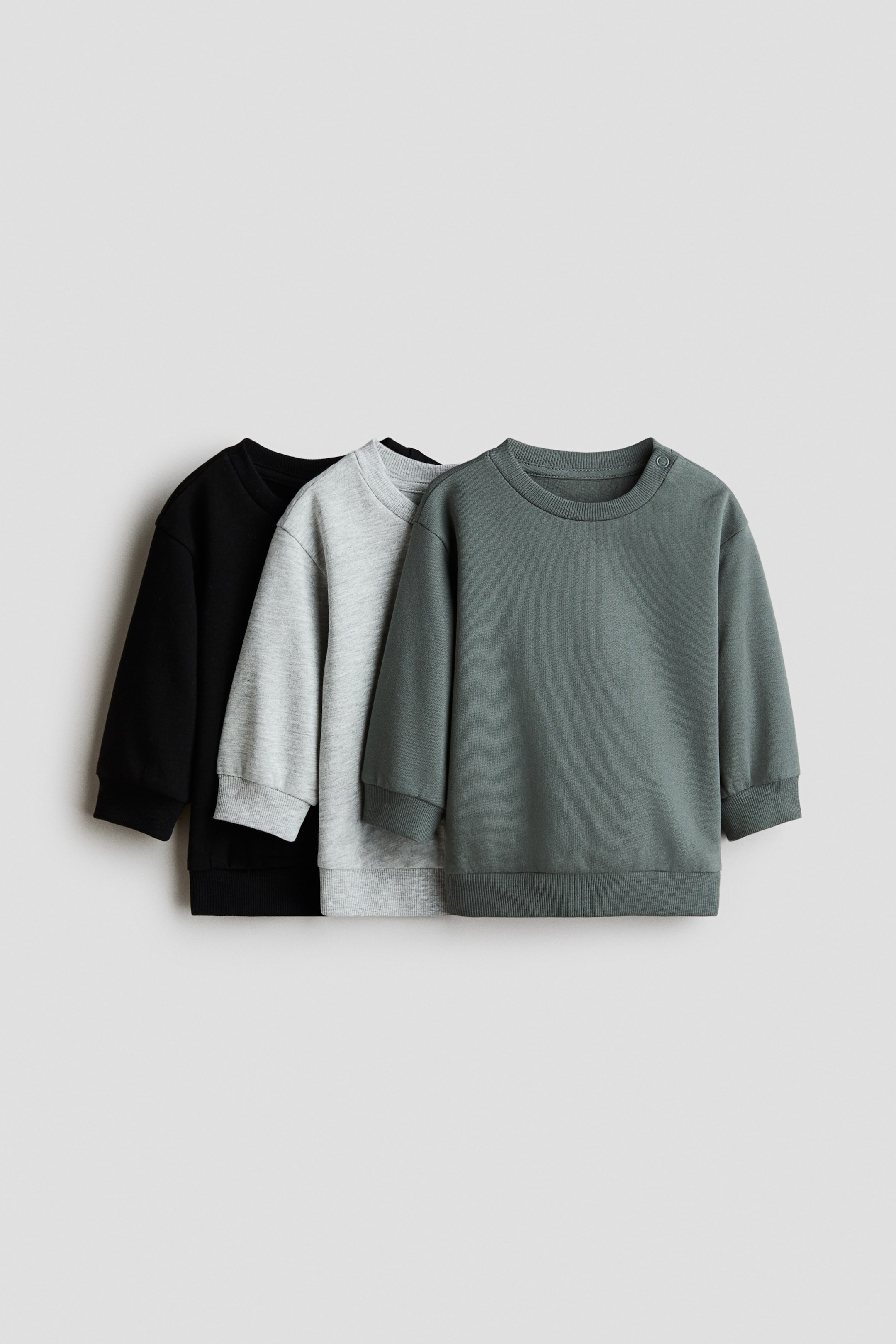 Se større billede: Et tre-pak af bomuldssweatshirts vises fra en frontvinkel, der viser en sort, en lys gråmeleret og en mørkegrøn sweatshirt. Alle tre har en rund halsudskæring, lange ærmer med ribmanchetter og en ribkant, mens den mørkegrønne sweatshirt også har trykknapper på venstre skulder.
