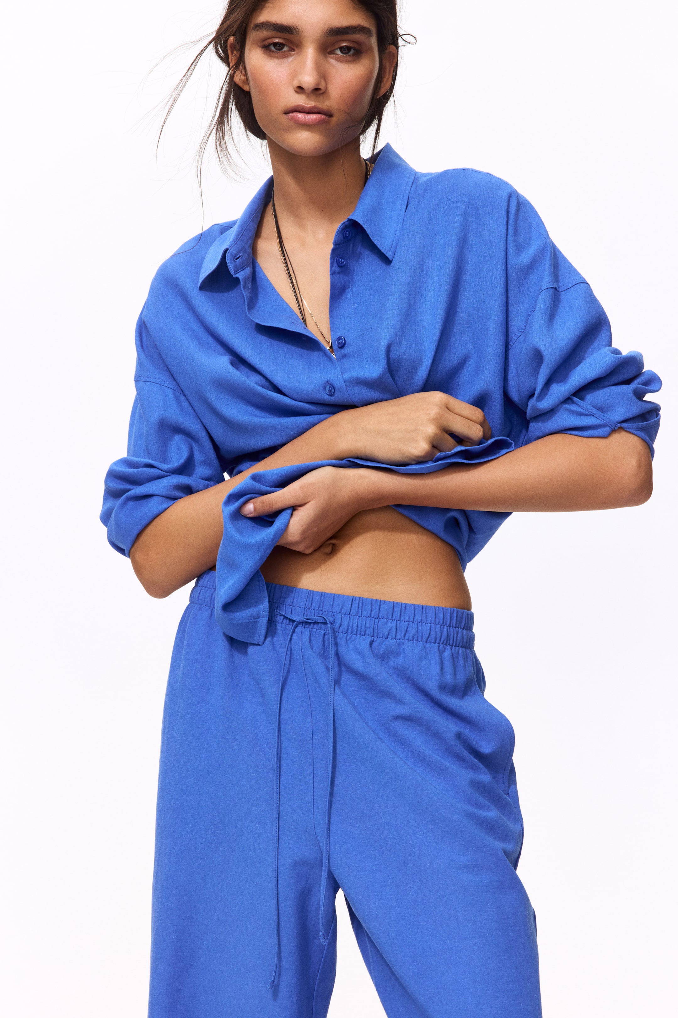 View larger image: The model is showcasing blue loose-fit trousers, characterized by an elasticated, drawstring waist, subtle side pockets, and wide legs. She is lifting a matching blue shirt, subtly revealing the waistline of the trousers.