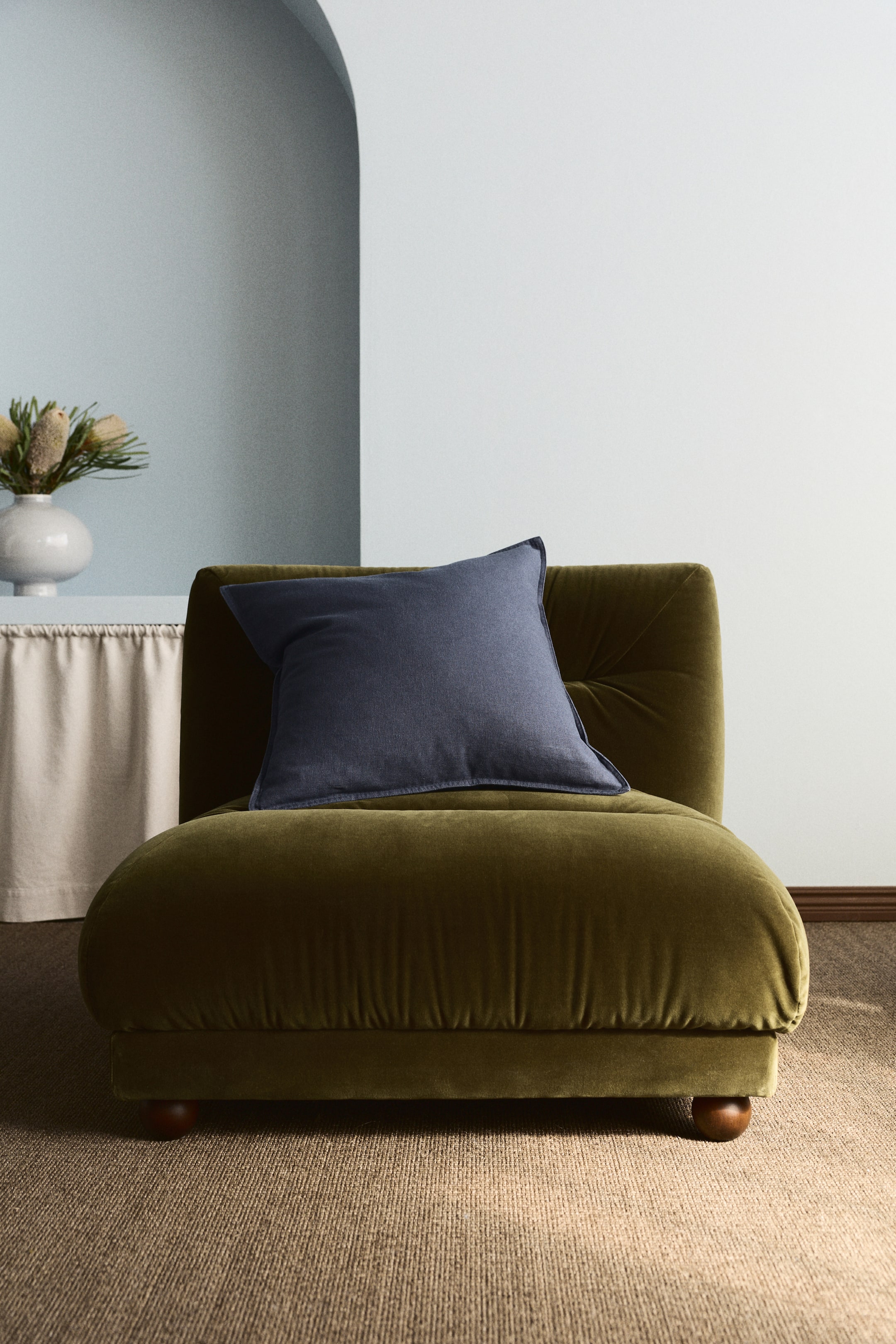 View larger image: An olive green velvet low lounge chair is positioned slightly to the right, featuring a tufted backrest, a rounded silhouette, and small wooden bun feet. A navy blue square cushion is placed diagonally on its seat.