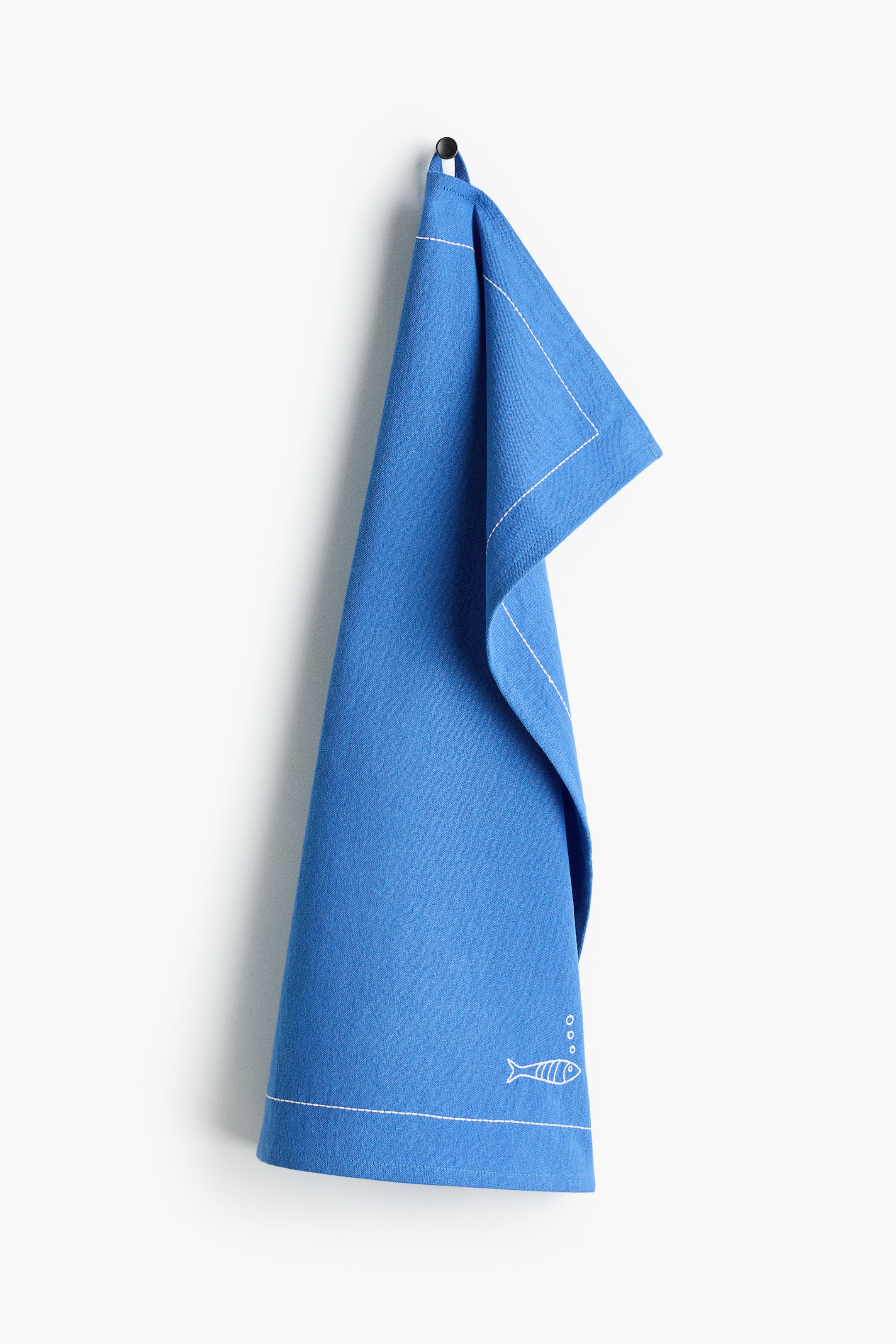Visualizza immagine più grande: Un canovaccio blu è appeso piegato a un gancio nero. Presenta cuciture ricamate bianche lungo i bordi e un piccolo motivo a pesce con bolle nell'angolo in basso a destra, e un'asola per appendere si forma su un lato corto.