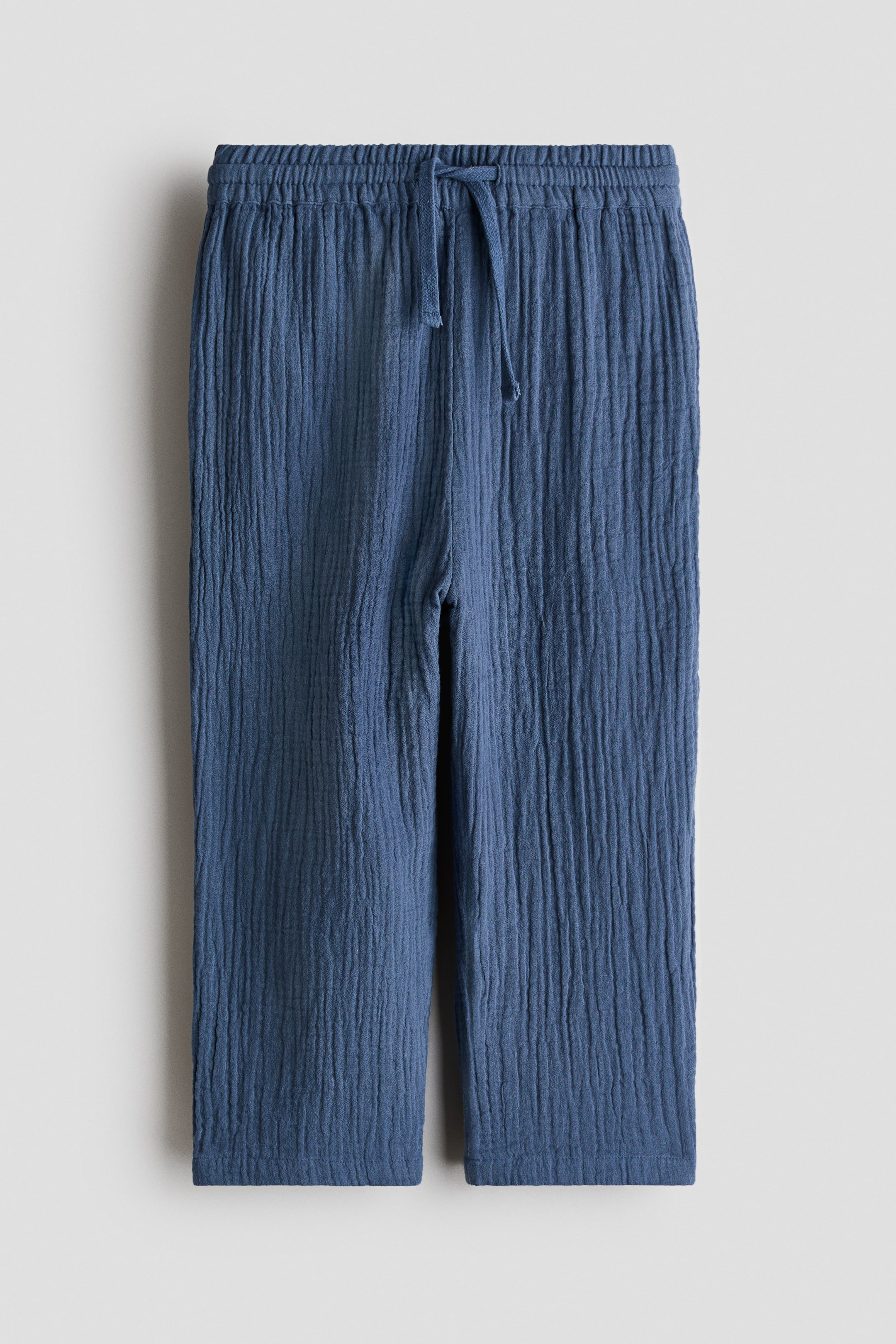 View larger image: A pair of blue crinkled trousers showcases an elasticated waistband with a central drawstring tie. The trousers feature a straight-leg cut and a textured vertical pattern throughout the fabric.