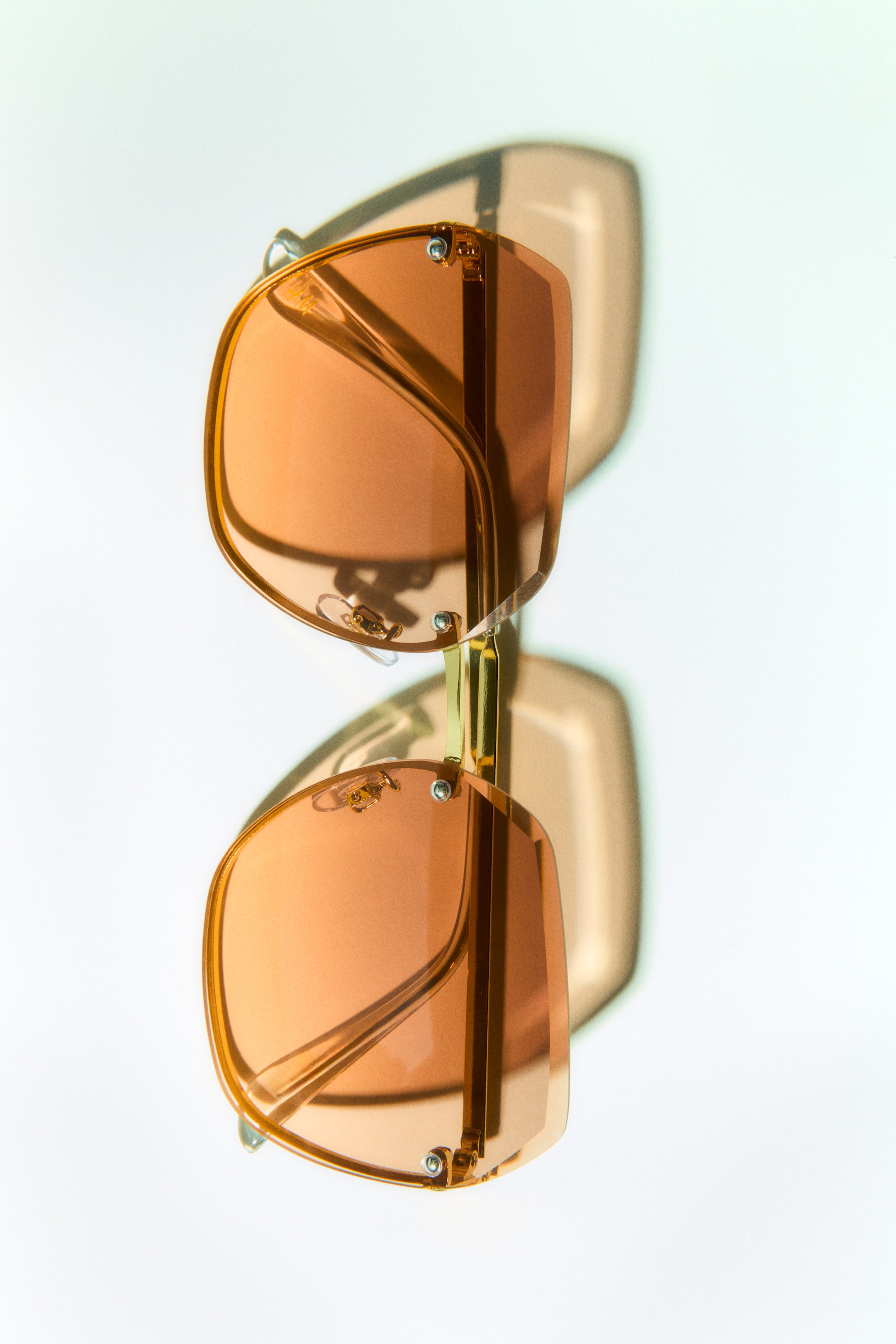 View larger image: Square sunglasses featuring hidden frames and metallic sidepieces. They showcase adjustable nose pads and tinted, amber-orange, UV-protective plastic lenses, facing front.