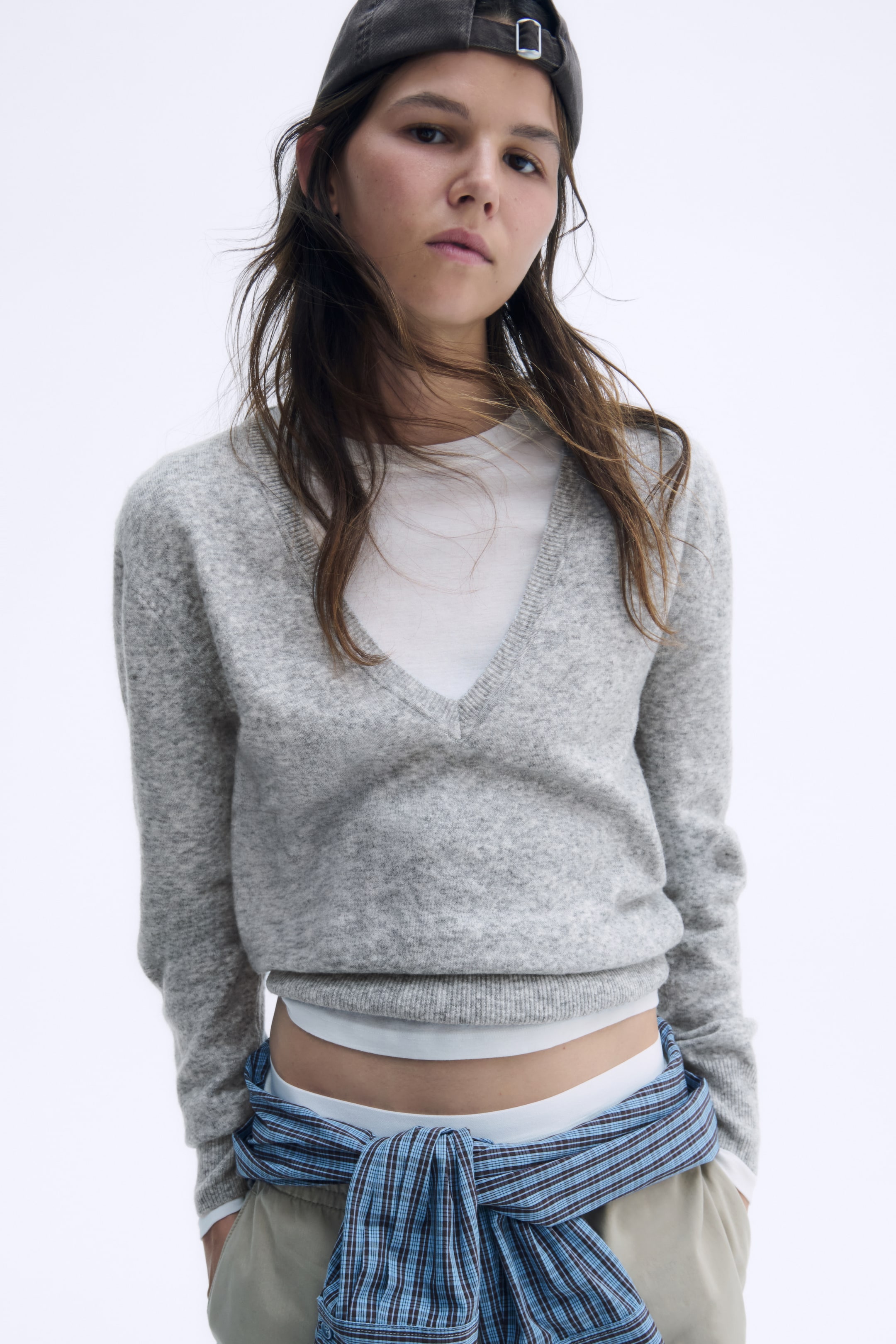 Grösseres Bild ansehen: Ein hellgrauer Strickpullover ist von vorne zu sehen und verfügt über einen tiefen, gerippt eingefassten V-Ausschnitt, der über einem weissen T-Shirt getragen wird. Der Pullover zeigt überschnittene Schultern, lange Ärmel mit gerippten Bündchen und einen gerippten Saum.
