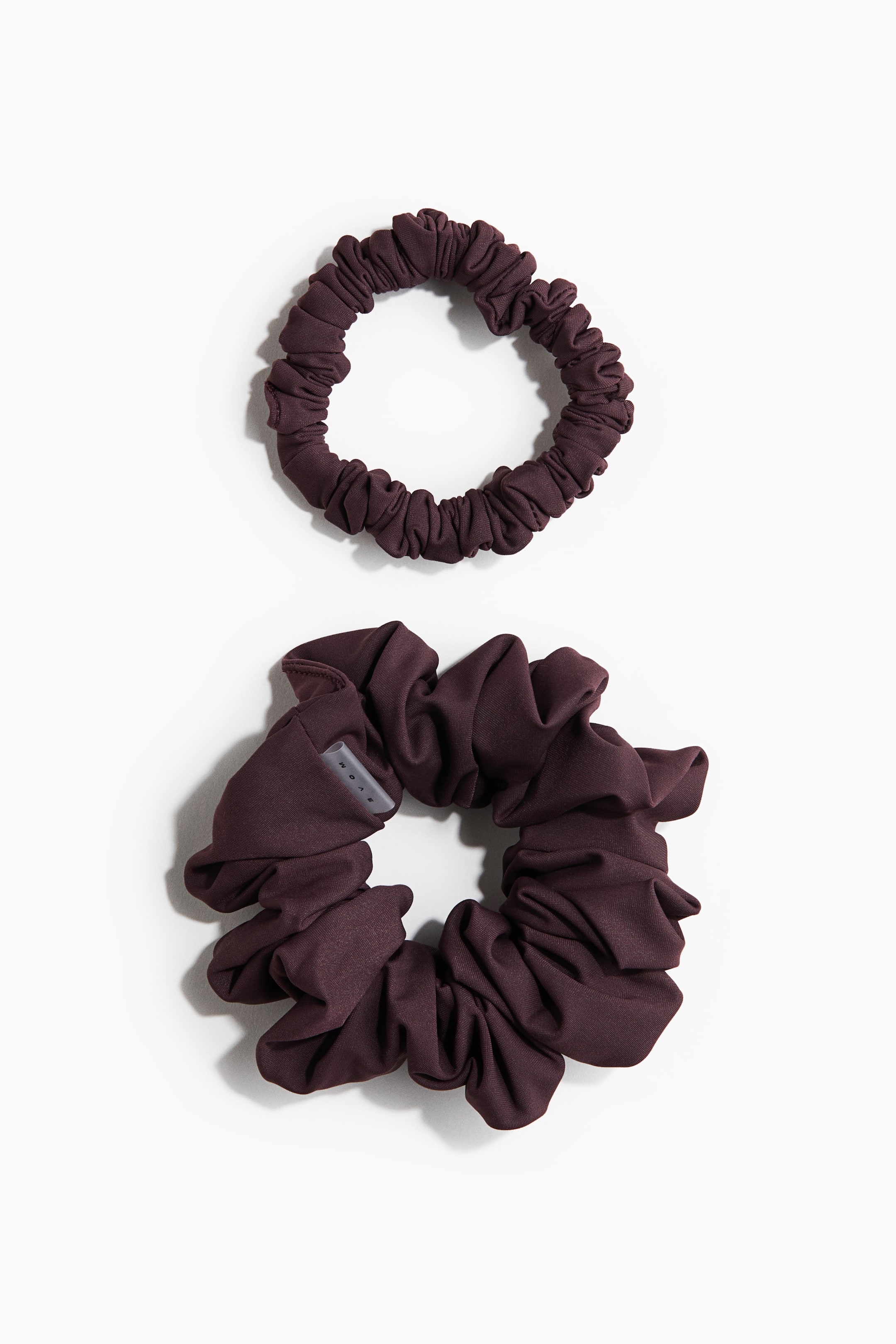 Visa större bild: Två scrunchies är mörklila och ligger plant. Den mindre scrunchien är tätt rynkad, medan den större har ett mer voluminöst, löst samlat tyg med en liten grå "WOAE"-etikett synlig på sidan.