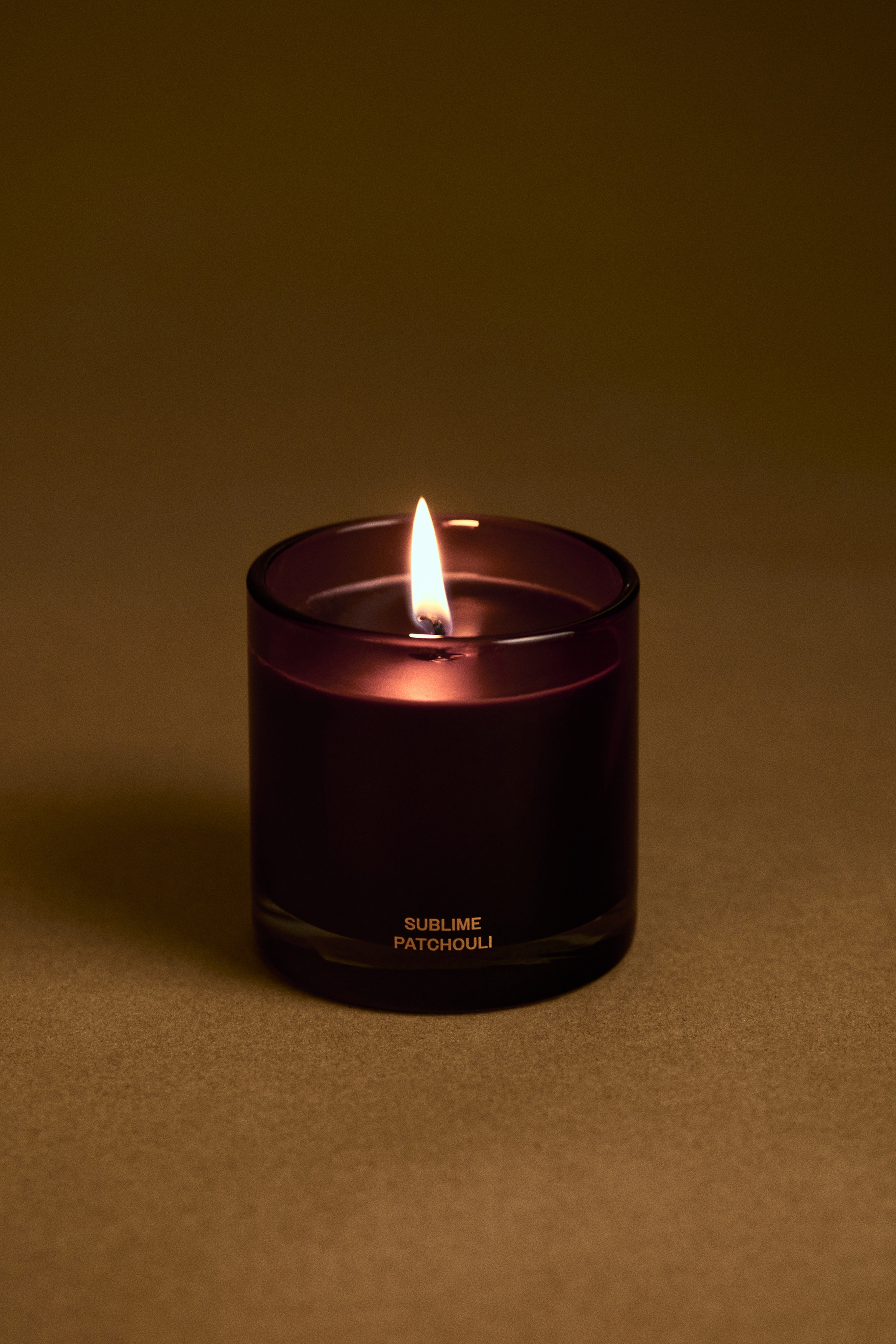 A scented candle in a cylindrical, translucent dark purple glass jar is standing facing the front, showcasing dark wax and a single central wick. The jar features a thick clear glass base and gold lettering near the bottom reading "SUBLIME PATCHOULI."