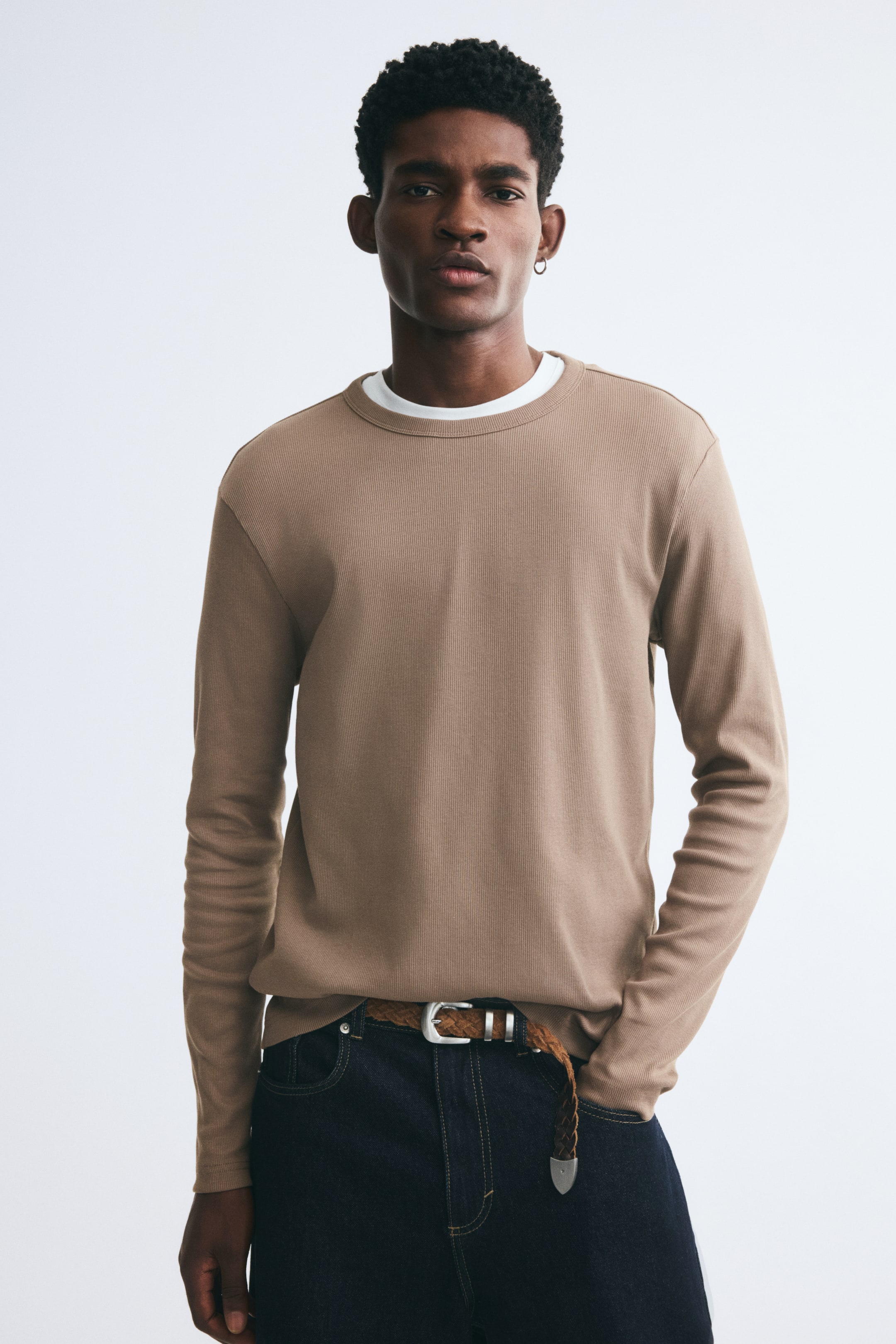 View larger image: A long-sleeve tee in light brown ribbed cotton jersey is being worn by the model, layered over a white crew-neck collar. The slim-fit tee features a round neckline and is styled with dark denim jeans and a braided belt.