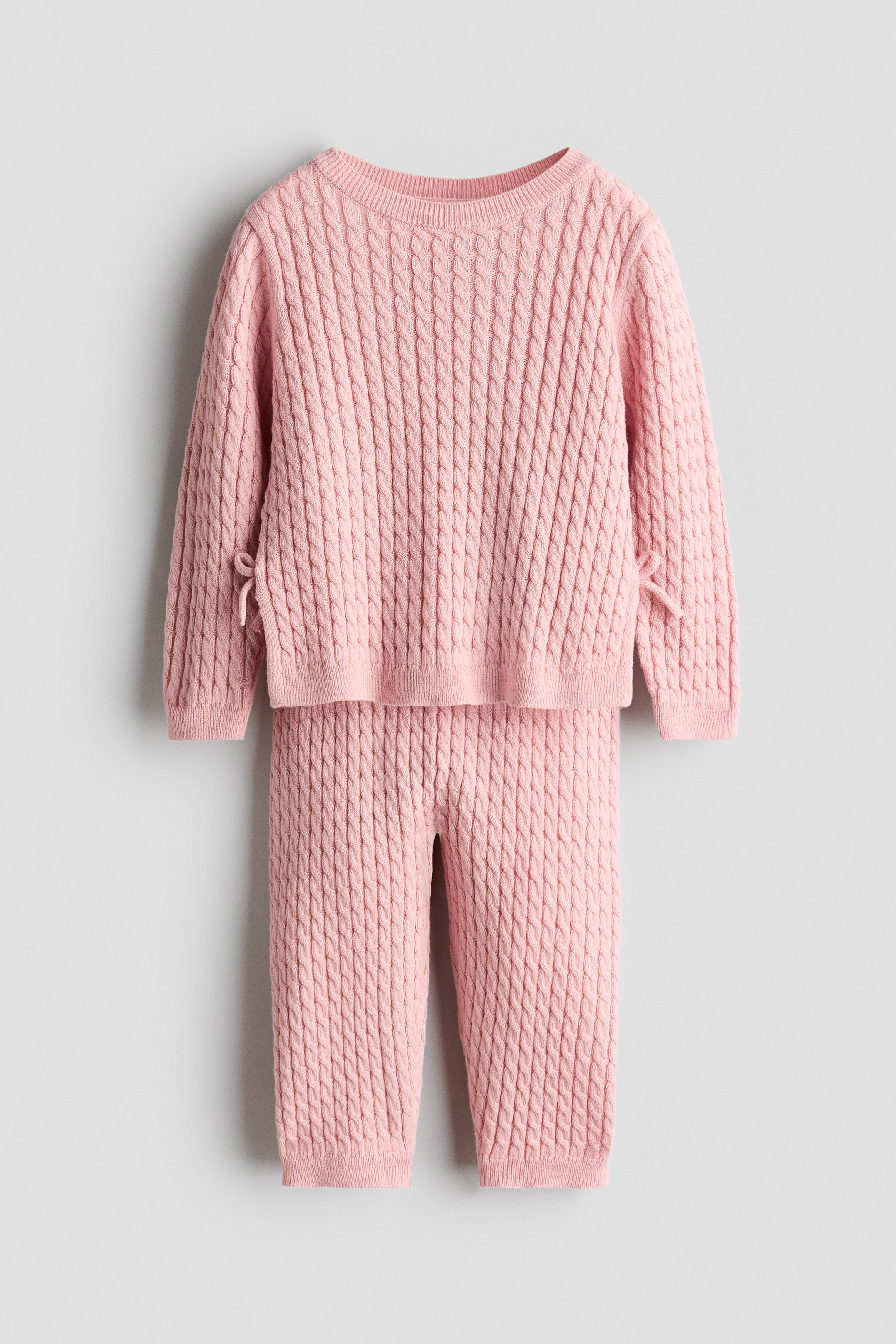 Visualizza l'immagine ingrandita: Un set composto da maglione e pantaloni in maglia a trecce rosa è in primo piano. Il maglione presenta uno scollo rotondo a coste, maniche lunghe con polsini a coste e un orlo a coste con lacci laterali. I pantaloni coordinati mostrano un design a lunghezza intera con polsini alla caviglia a coste.