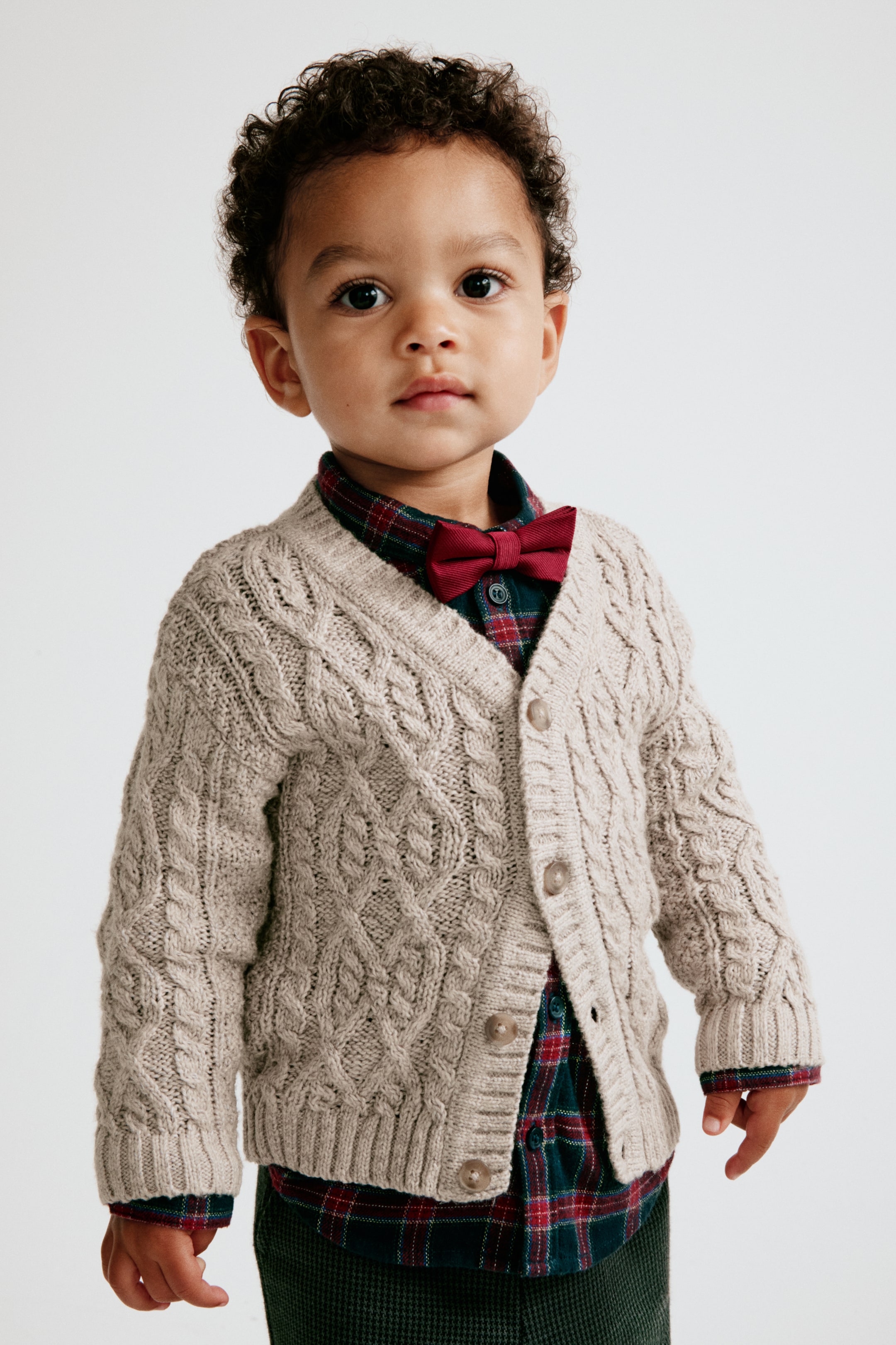 Ver imagem maior: Um cardigan bege em malha entrançada é apresentado de frente, exibindo um decote em V, abertura frontal com botões e punhos e bainha canelados. O cardigan é usado por cima de uma camisa aos quadrados com botões e uma gravata borboleta vermelha, destacando o seu ajuste estruturado, mas confortável.