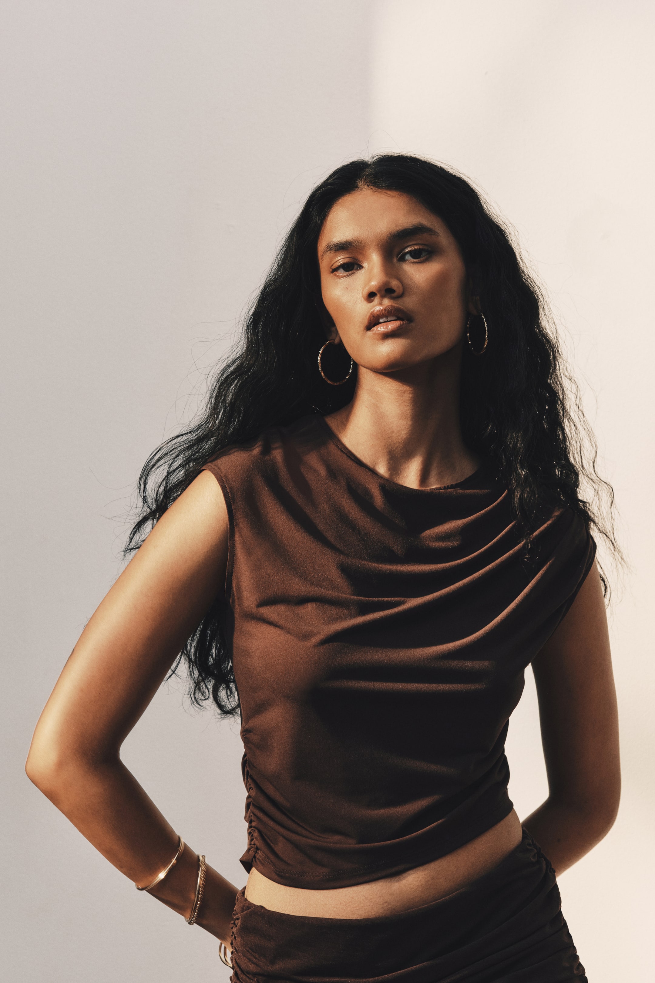 View larger image: A dark brown stretch jersey top featuring a round neckline, cap sleeves, and a draped bodice is worn by the model. The top features pleats on one shoulder and a gathered side seam, creating an asymmetrical cropped hem styled with matching draped bottoms.
