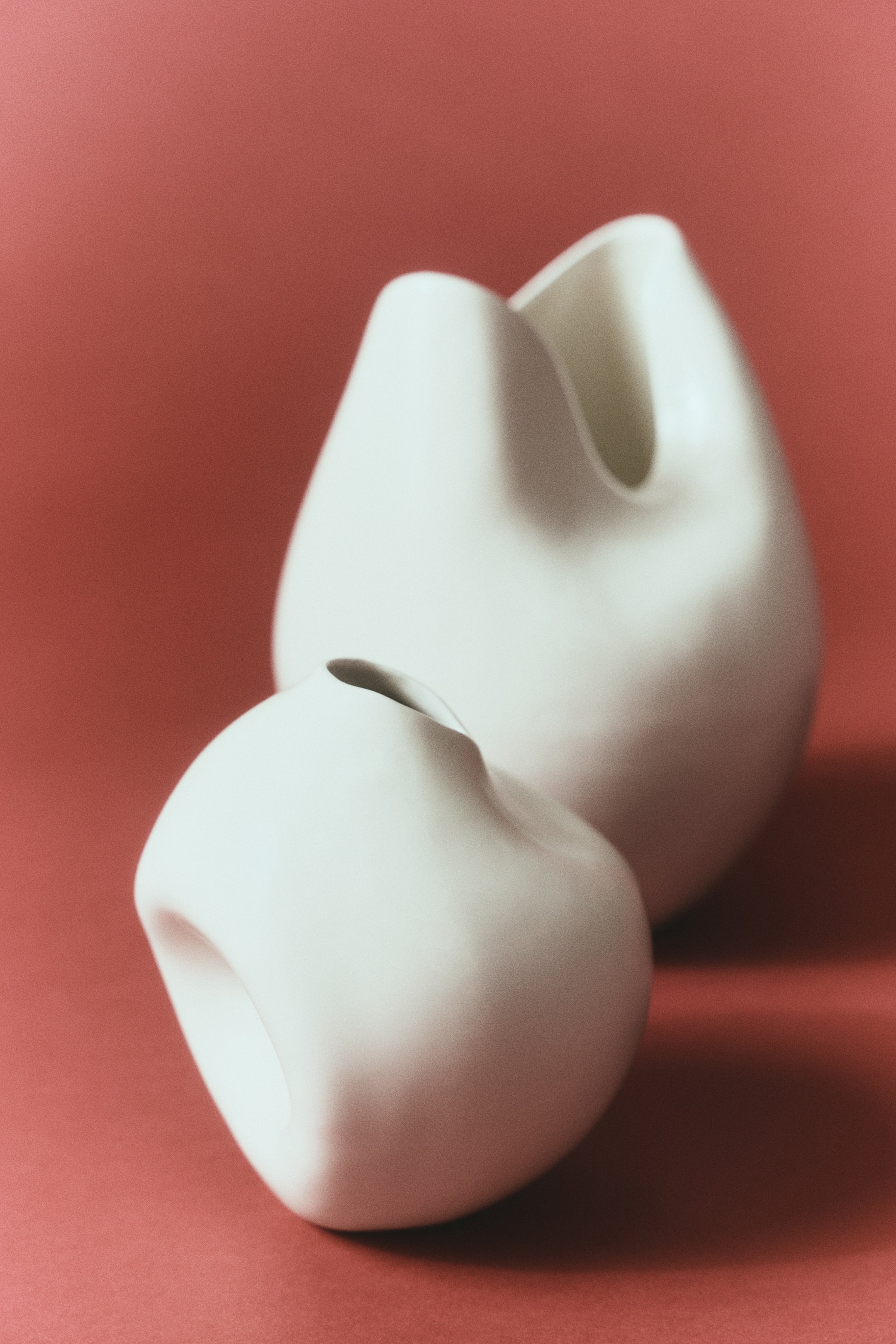 Visualizza immagine più grande: Due vasi in ceramica bianco sporco mostrano design asimmetrici e scultorei. Il vaso più piccolo in primo piano presenta una forma arrotondata e ორგანica con un’apertura stretta, mentre il vaso più grande sullo sfondo presenta una forma più alta e bulbosa con un bordo ampio e ondulato.