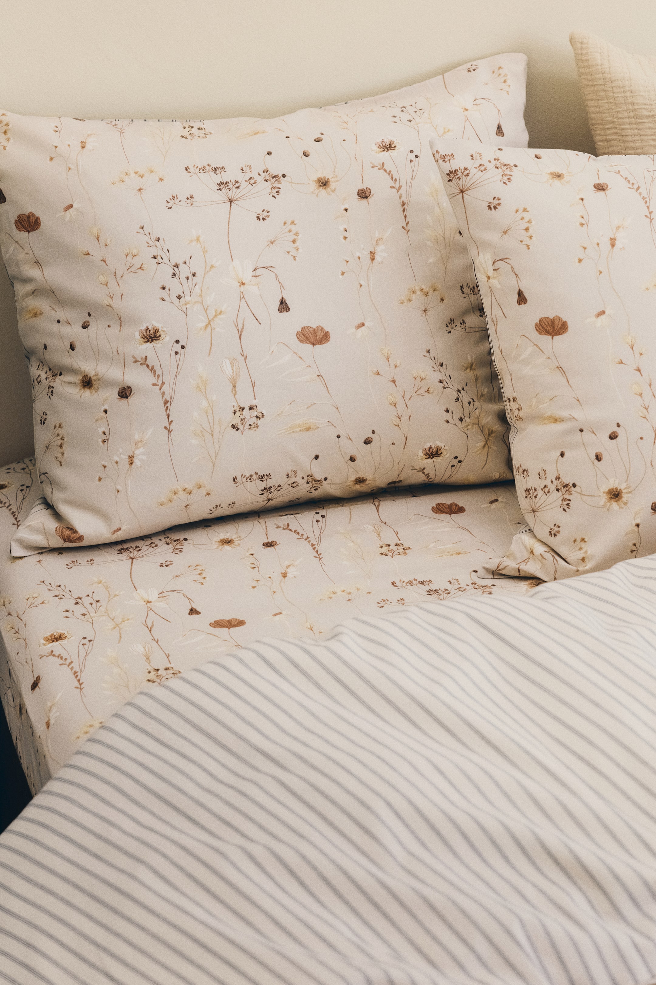 Ver imagem maior: Um lençol ajustado exibe um padrão delicado de flores silvestres e ervas dispersas em tons suaves de castanho, creme e amarelo sobre um fundo bege claro, com um edredão às riscas a cobri-lo parcialmente.