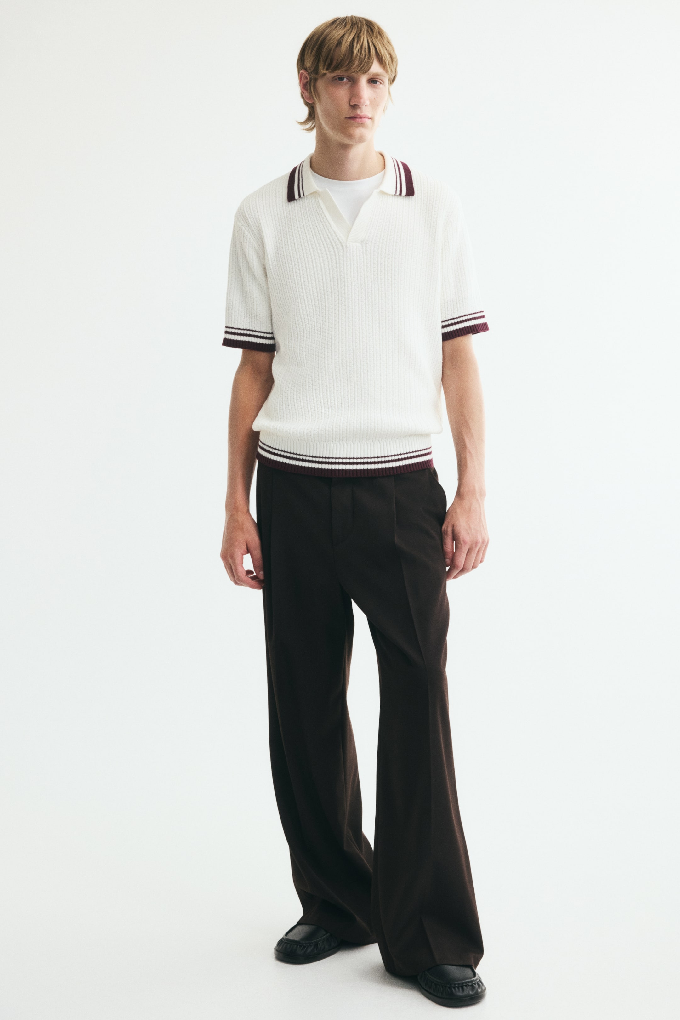 View larger image: A cream-coloured, textured knit polo shirt is worn by the model, featuring a ribbed open collar, cuffs, and hem with contrasting burgundy and white striped trims. The short-sleeve, regular-fit shirt is layered over a white crew-neck T-shirt and paired with dark brown wide-leg trousers.