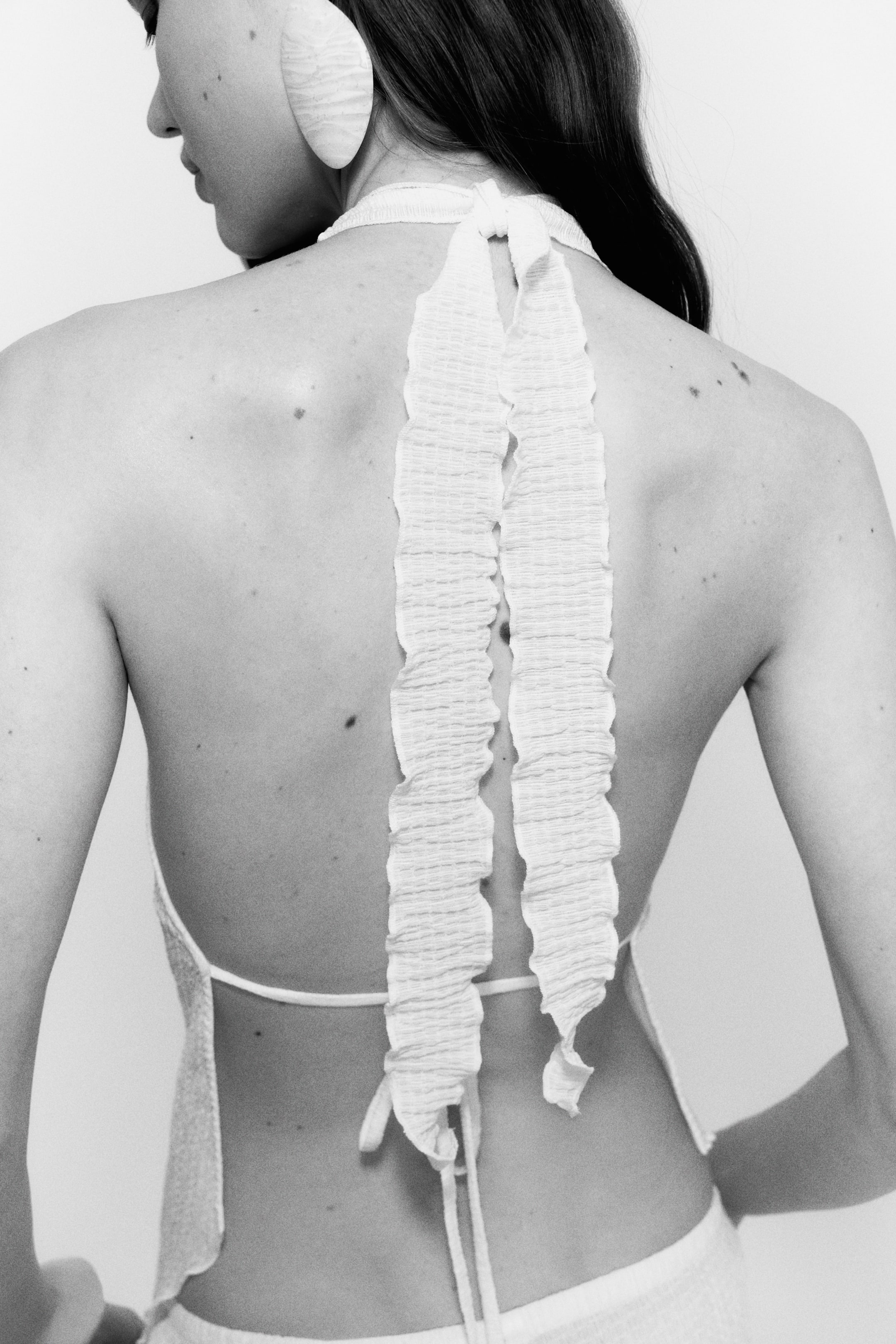 Größeres Bild anzeigen: Ein helles Neckholder-Top mit zwei vertikalen, gekräuselten Trägern, die über den offenen Rücken verlaufen und am Nacken sowie am unteren Rücken gebunden werden.