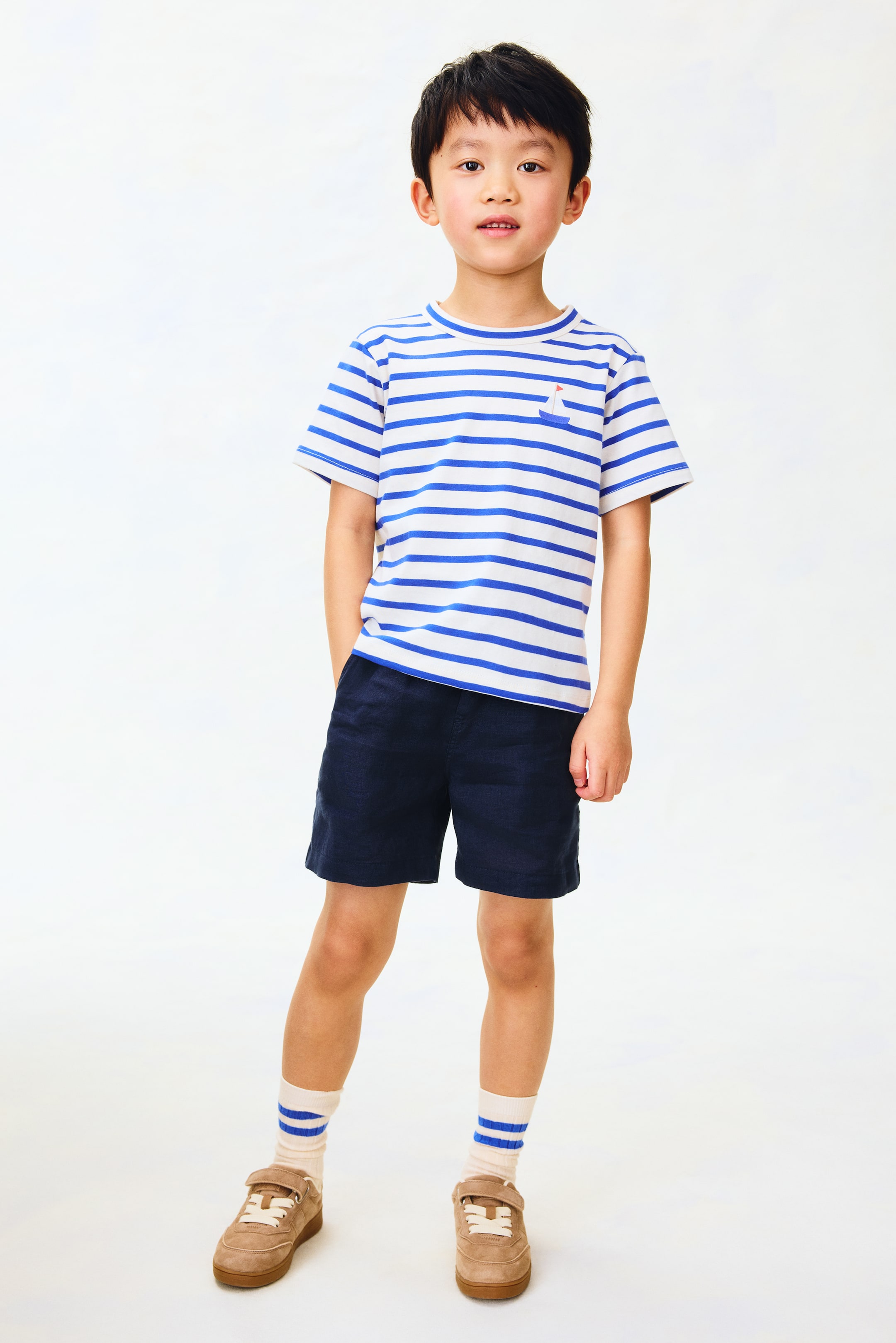 View larger image: A white T-shirt featuring blue horizontal stripes, a round neckline, and short sleeves, with a small red and white sailboat emblem adorning the left chest. Dark blue shorts, reaching above the knees, complete the outfit, paired with cream-colored crew socks displaying two blue stripes, and tan low-top sneakers featuring white laces and a single Velcro strap closure.