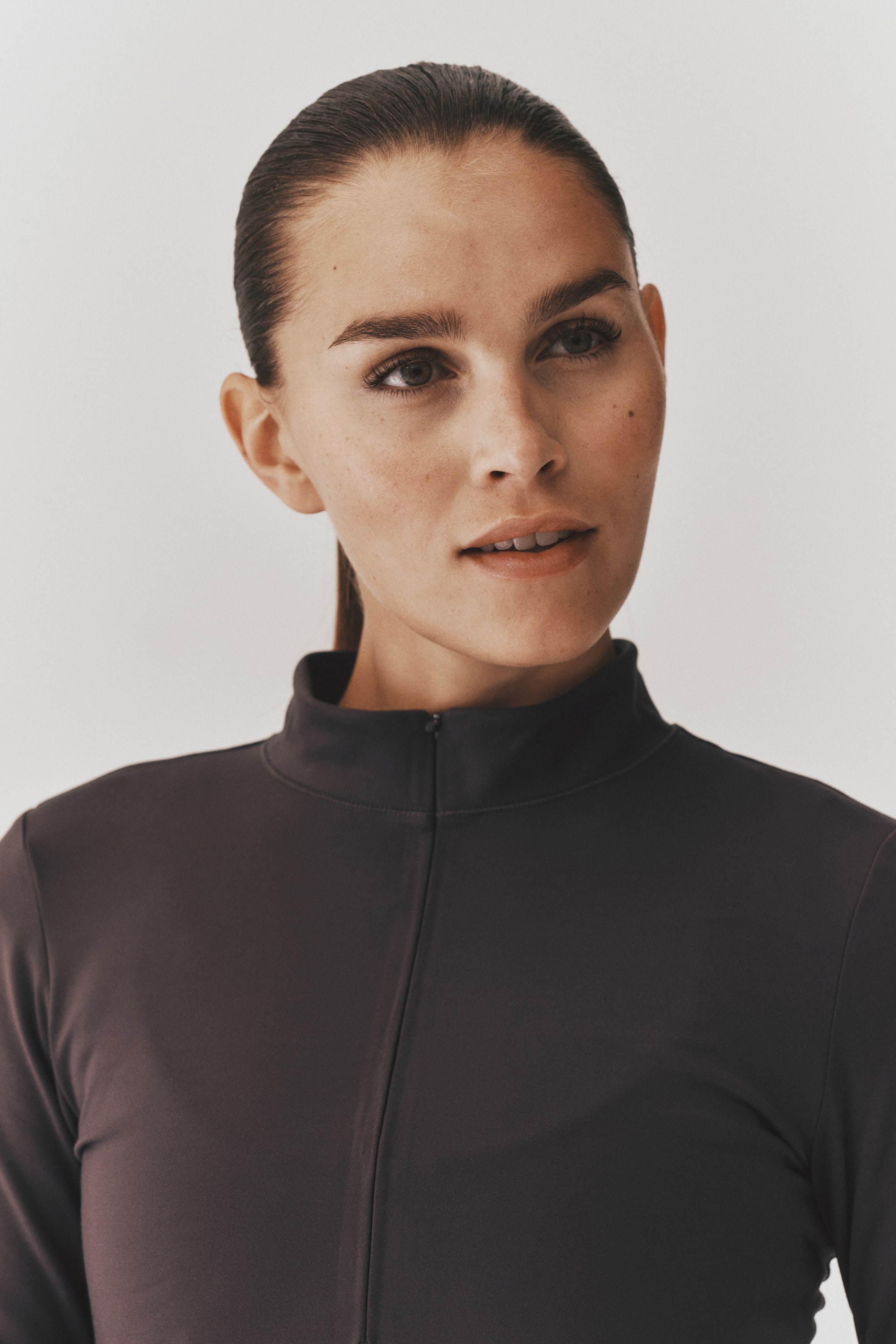 View larger image: A dark brown, long-sleeved activewear top is being worn by the model. The top features a fitted silhouette, a mock-neck collar, and a central front zipper closure.