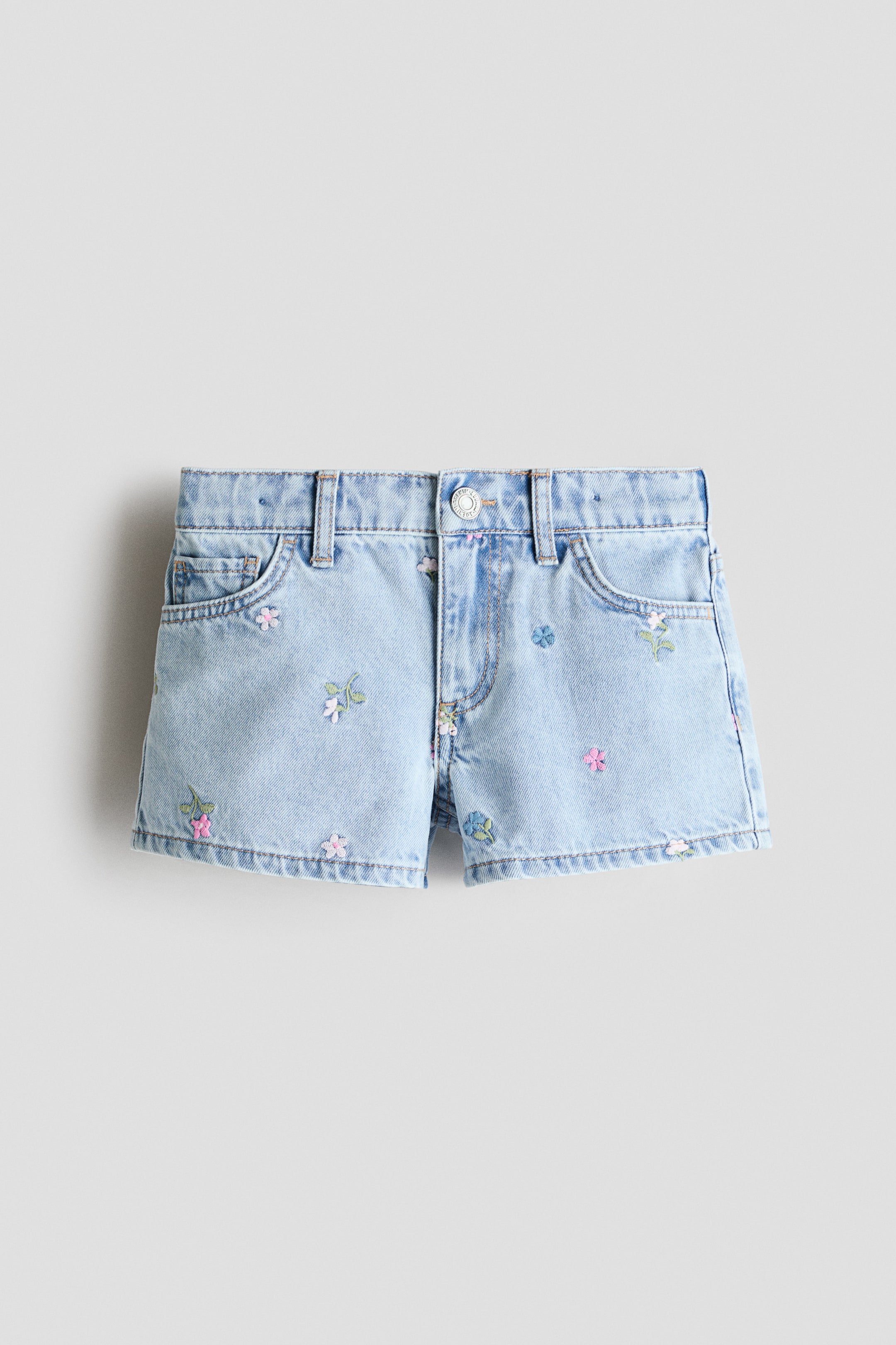 View larger image: The 5-pocket shorts in light blue cotton denim are shown from the front, showcasing an adjustable, elasticated waist with belt loops and a zip fly fastened by a press-stud. Scattered floral embroidery in pink, green, blue, and white is visible across the fabric.