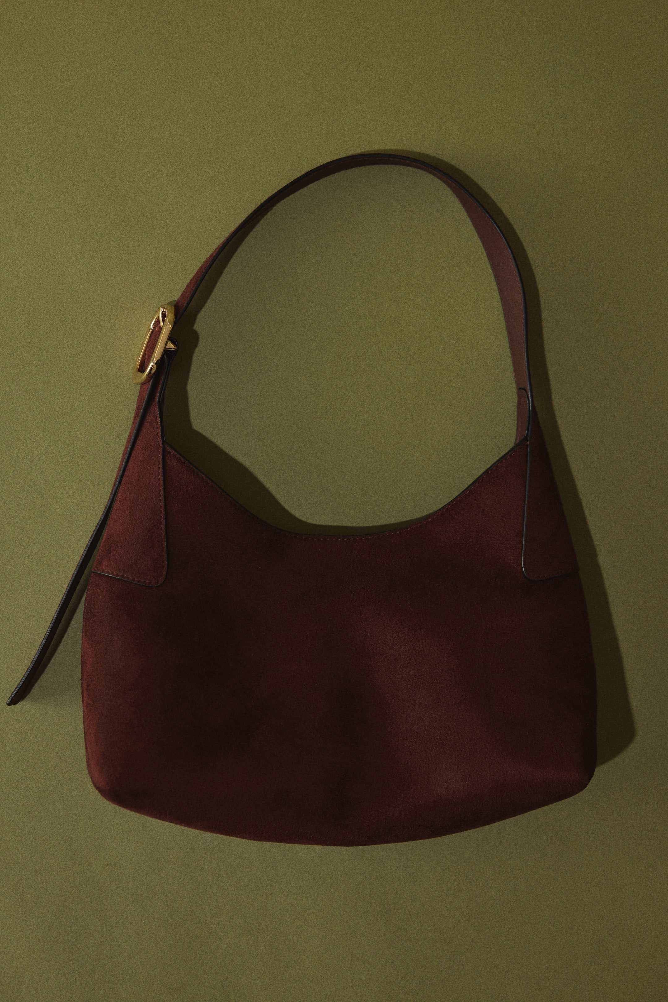 Größeres Bild ansehen: Eine Hobo-Bag aus burgunderrotem Wildleder liegt flach und zeigt ihre weiche, halbmondförmige Silhouette. Ein dünner, dunkler Schulterriemen verläuft vom oberen Rand der Tasche und ist auf der linken Seite mit einer goldfarbenen Schnalle befestigt.