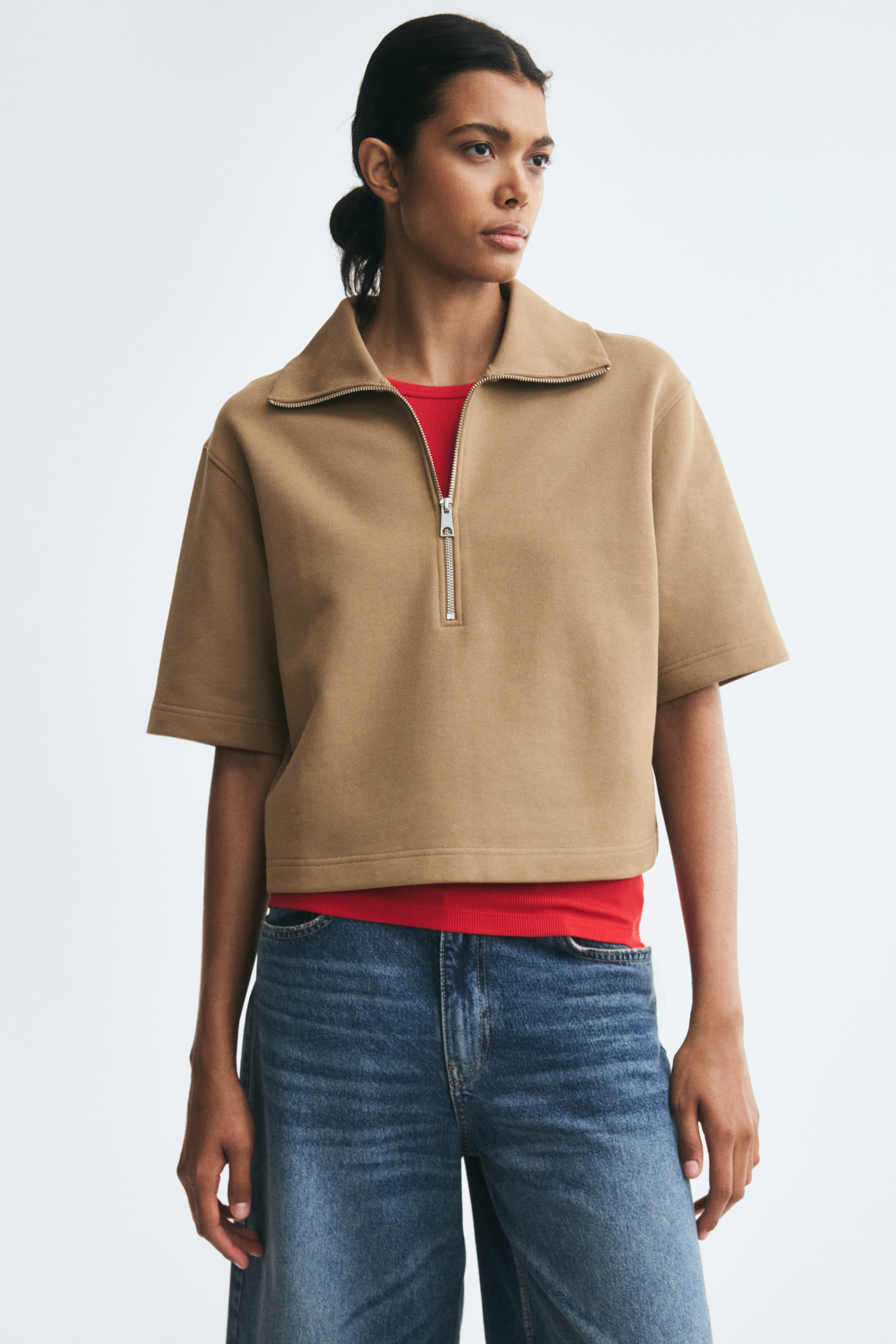 View larger image: A beige polo shirt in lightweight sweat fabric is being worn by the model, layered over a red top and paired with blue jeans. The shirt features a collar with an open half-zip, dropped shoulders, and wide, elbow-length sleeves.
