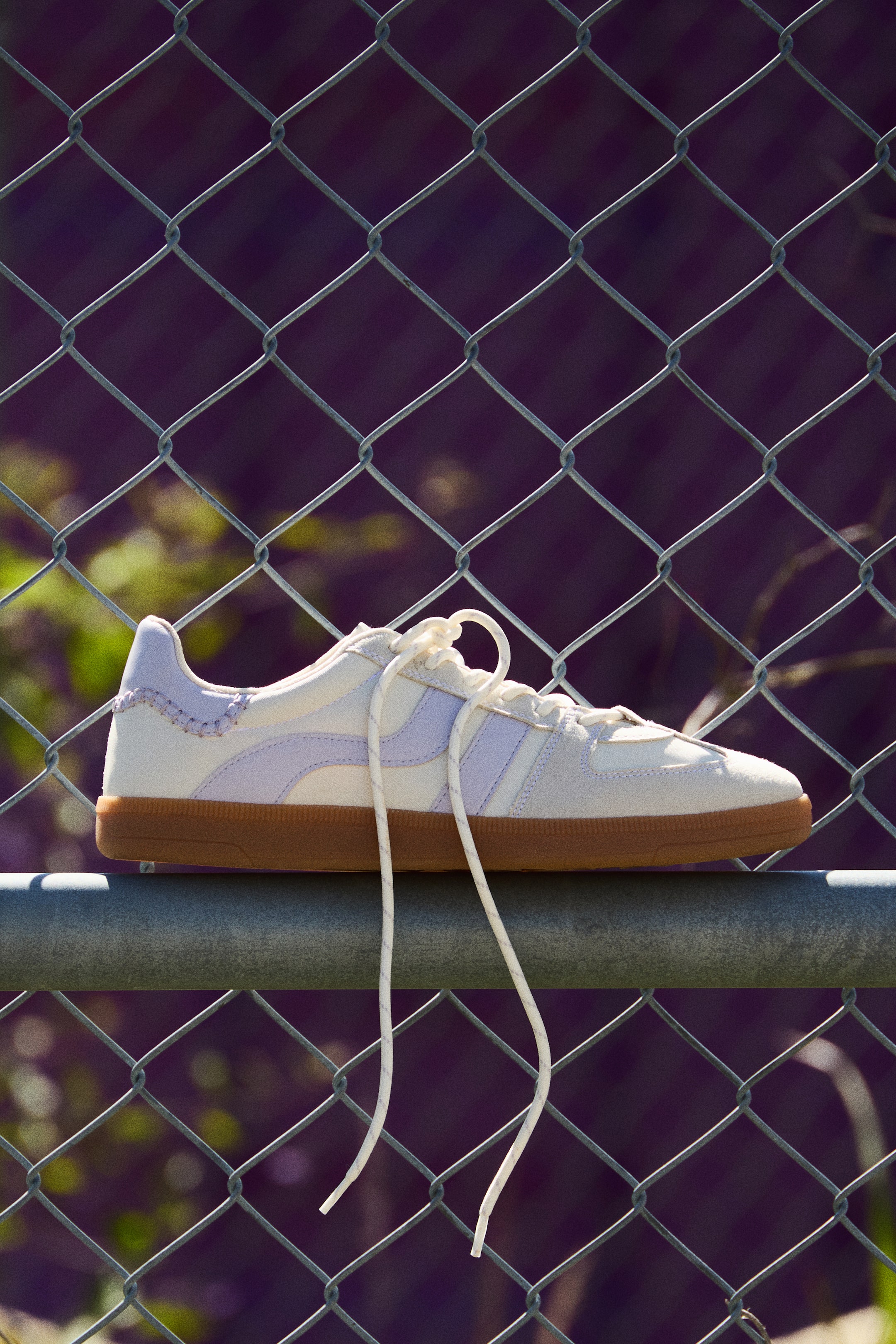 Vezi imaginea mai mare: Un pantof sport cu profil jos prezintă un material superior de culoare crem deschis, cu dungi laterale lavandă, margine superioară căptușită și șireturi albe desfăcute în față. Este așezat pe o talpă de gumă maro-roșiatică de 2,5 cm.