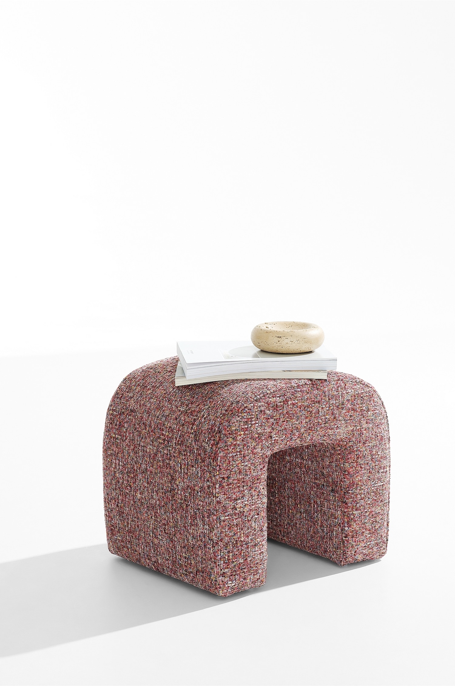 Un pouf a forma di U è posizionato con unâangolazione di tre quarti, mettendo in risalto il suo design ad arco con la parte superiore arrotondata e il centro cavo. Lo sgabello è interamente rivestito in un tessuto intrecciato testurizzato e multicolore, con sfumature di rosa, rosso, giallo e crema.