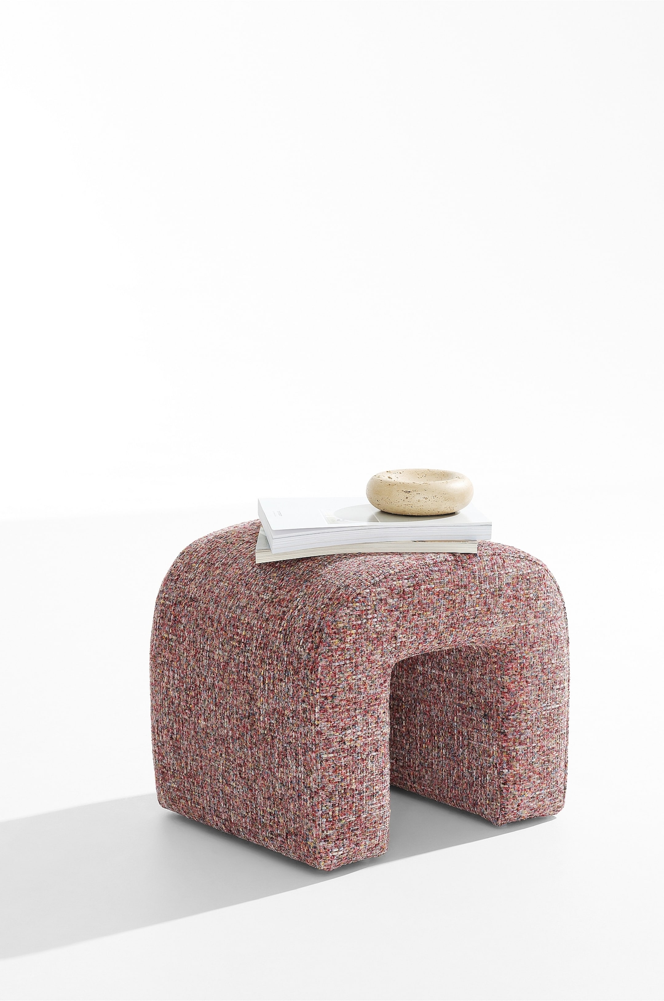 Visualizza immagine più grande: Un pouf a forma di U rivestito in un tessuto intrecciato materico, multicolore, nei toni del rosso e del rosa, è mostrato da un’angolazione a tre quarti. Due riviste e una piccola ciotola rotonda in pietra sono appoggiate sulla sua superficie superiore curva.