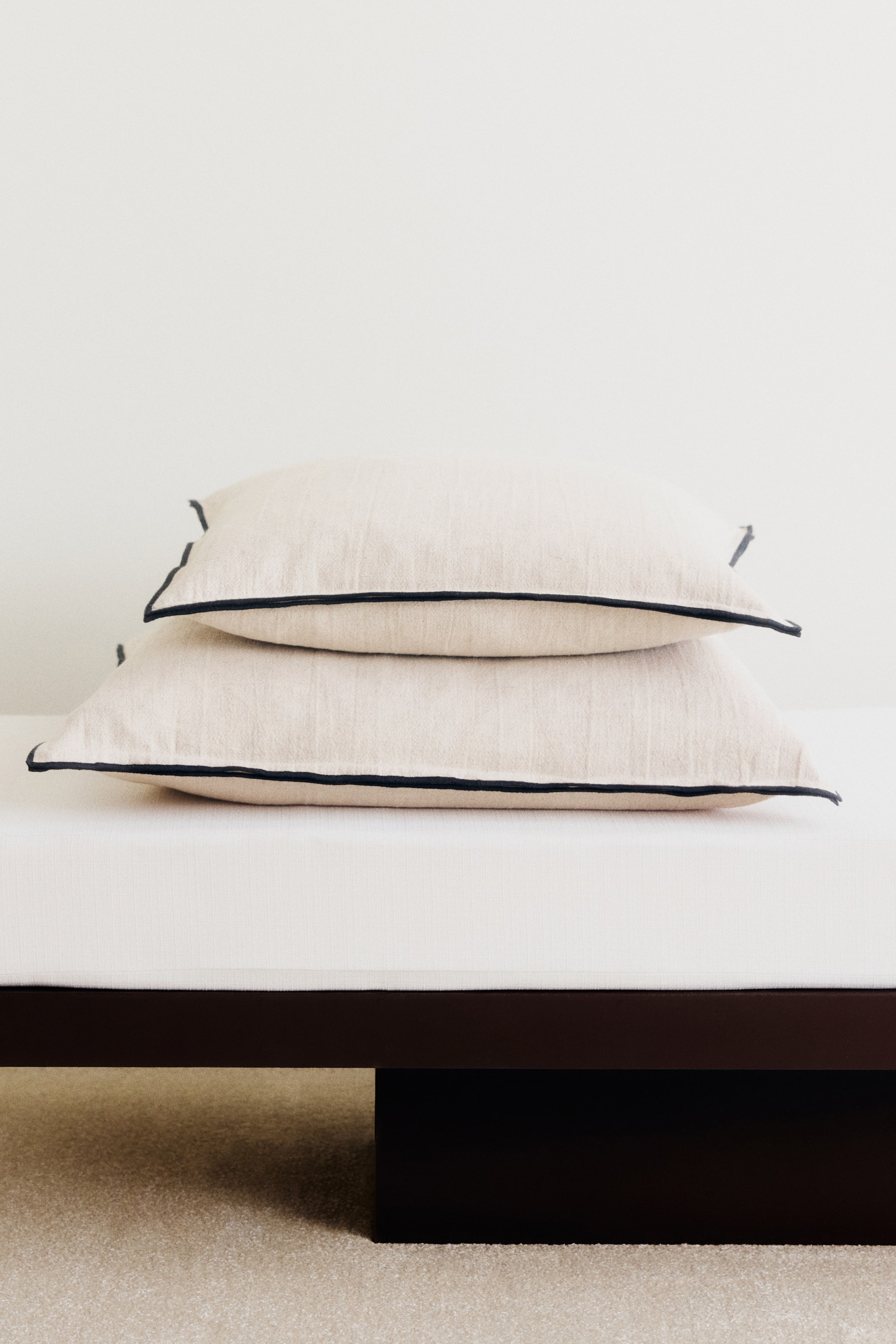 Agrandir l'image: Deux housses de coussin beige en mélange de lin et de coton texturé reposent l’une sur l’autre, présentées de face. Chaque housse met en valeur une double bordure surjetée contrastante de couleur foncée le long des bords.