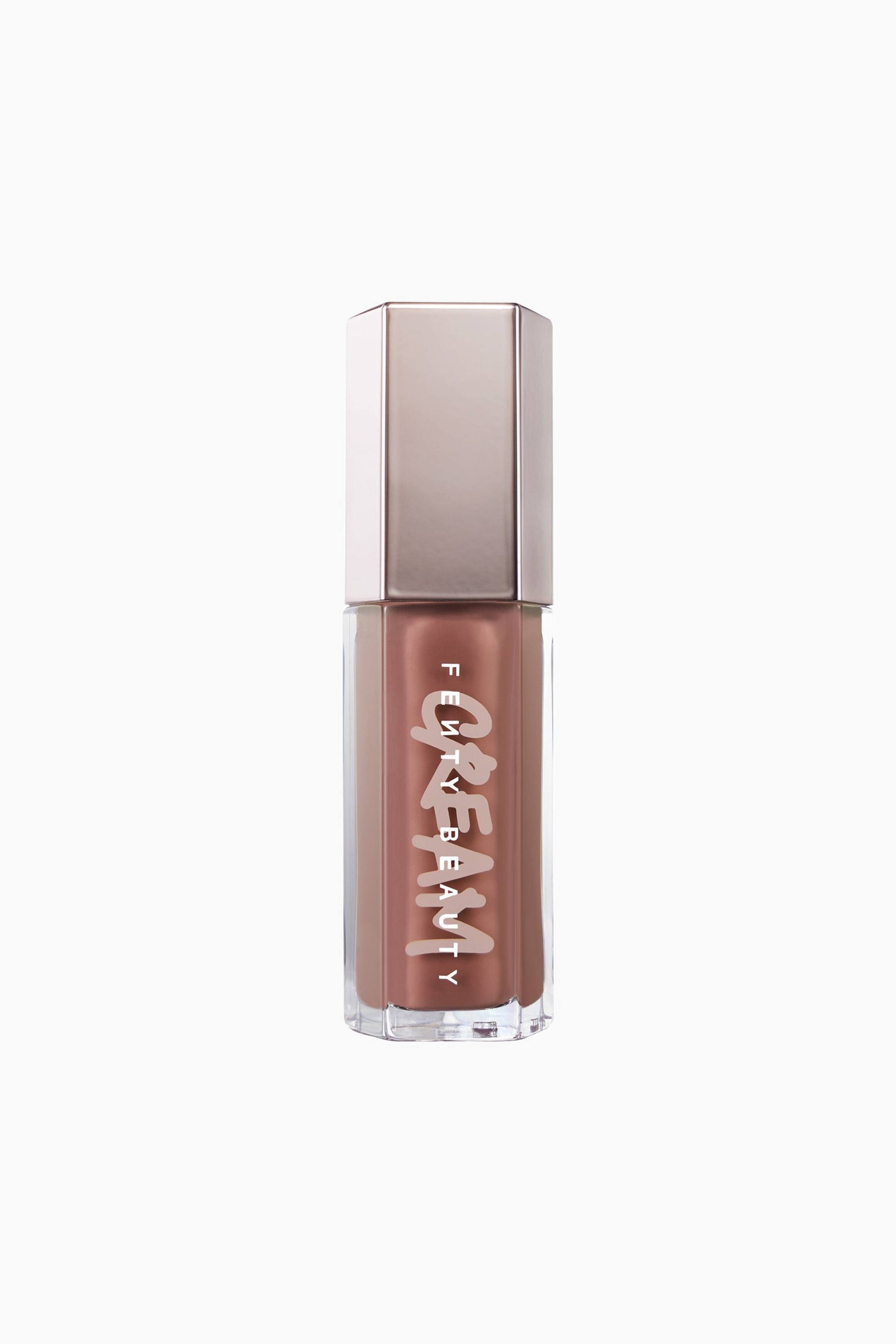 Näytä suurempi kuva: Fenty Beauty Gloss Bomb Cream -huulikiilto seisoo pystyssä esitellen kirkasta kuusikulmaista tuubia, joka on täytetty kermaisella, lämpimän sävyisellä ruskealla huulikiillolla. Tuubin päällä on heijastava metallinhohtoinen ruusukullanvärinen kuusikulmainen korkki, ja siinä on tyylitelty valkoinen sana "CREAM" sekä sen päällä pystysuunnassa painettu "FENTY BEAUTY".