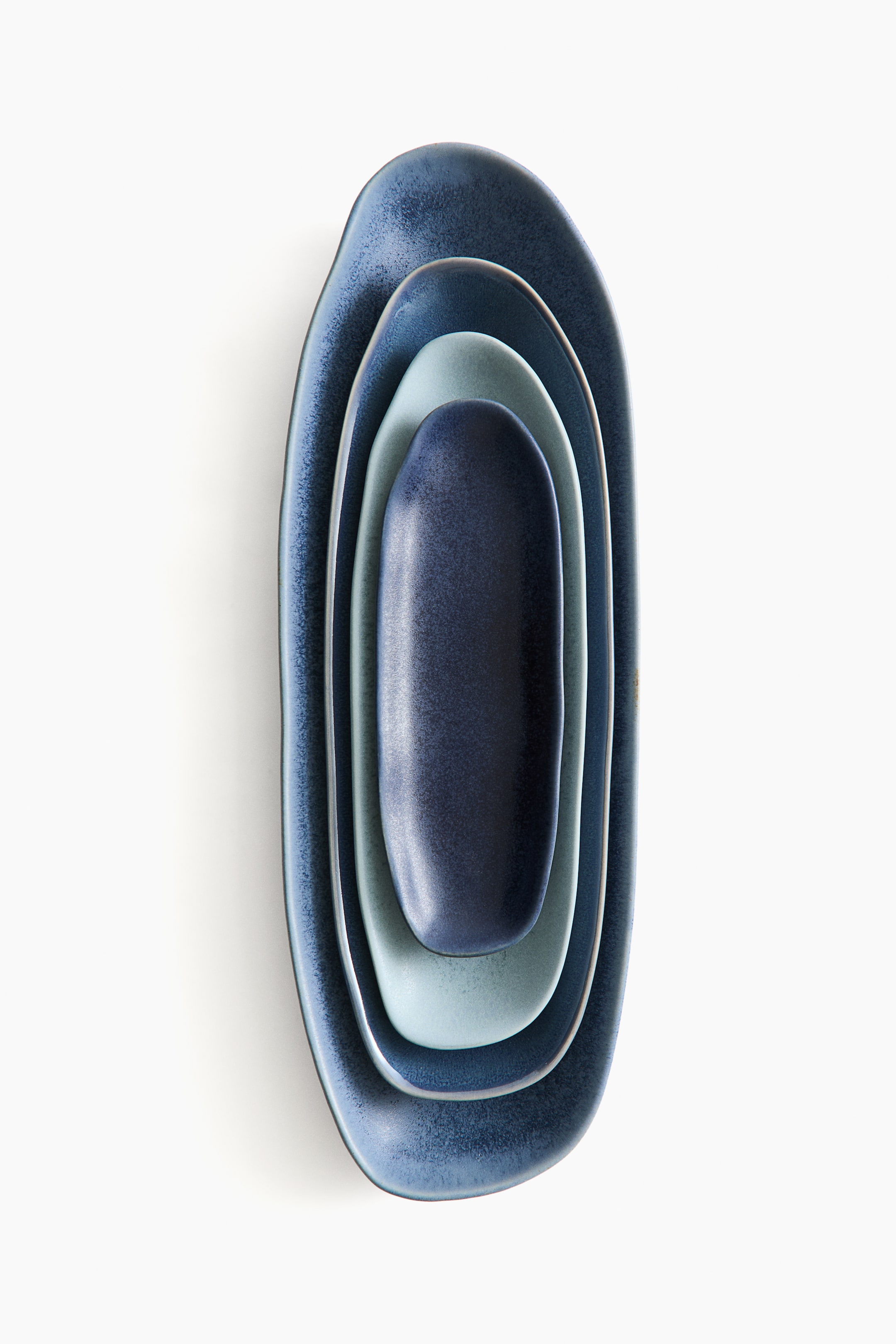 Agrandir l'image: Un ensemble de quatre plats de service oblongs en grès est emboîté du plus grand au plus petit, orienté vers l’avant dans une composition verticale. Les plats mettent en valeur des émaux texturés uniques dans différentes nuances de bleu, allant du bleu marine profond au bleu moyen moucheté et au bleu glace pâle.