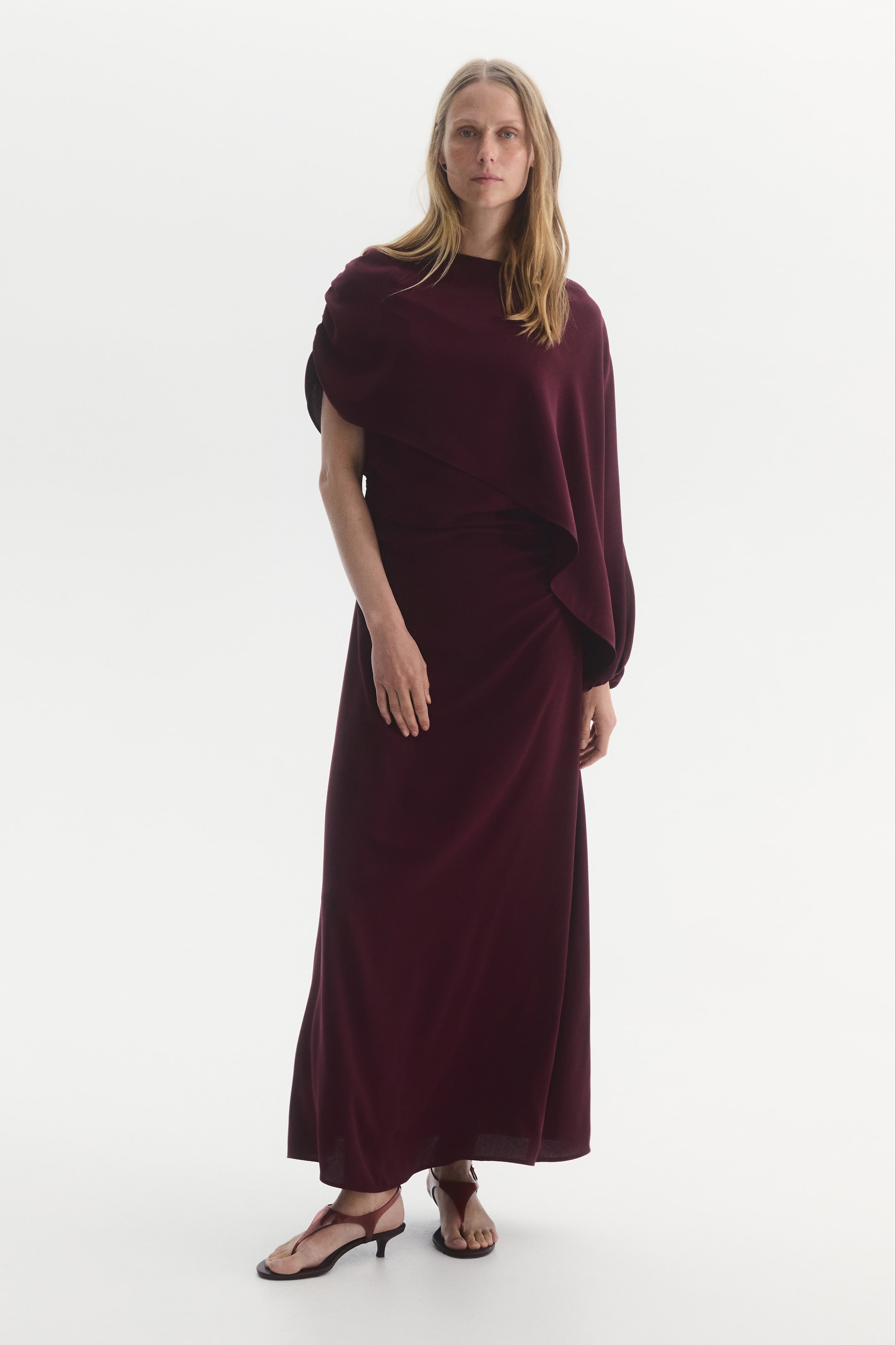 Näytä suurempi kuva: Malli esittelee MAMA Cape Detail Nursing Dress -mekkoa syvän viininpunaisessa sävyssä, korostaen sen maksimittapituista siluettia ja korkeaa pääntietä. Mekossa on epäsymmetrinen, laskeutuva viittamainen päällysosa, joka peittää toisen olkapään ja kulkee rinnan yli. Asuun on yhdistetty sävyyn sopivat viininpunaiset remmisandaalit matalalla korolla.