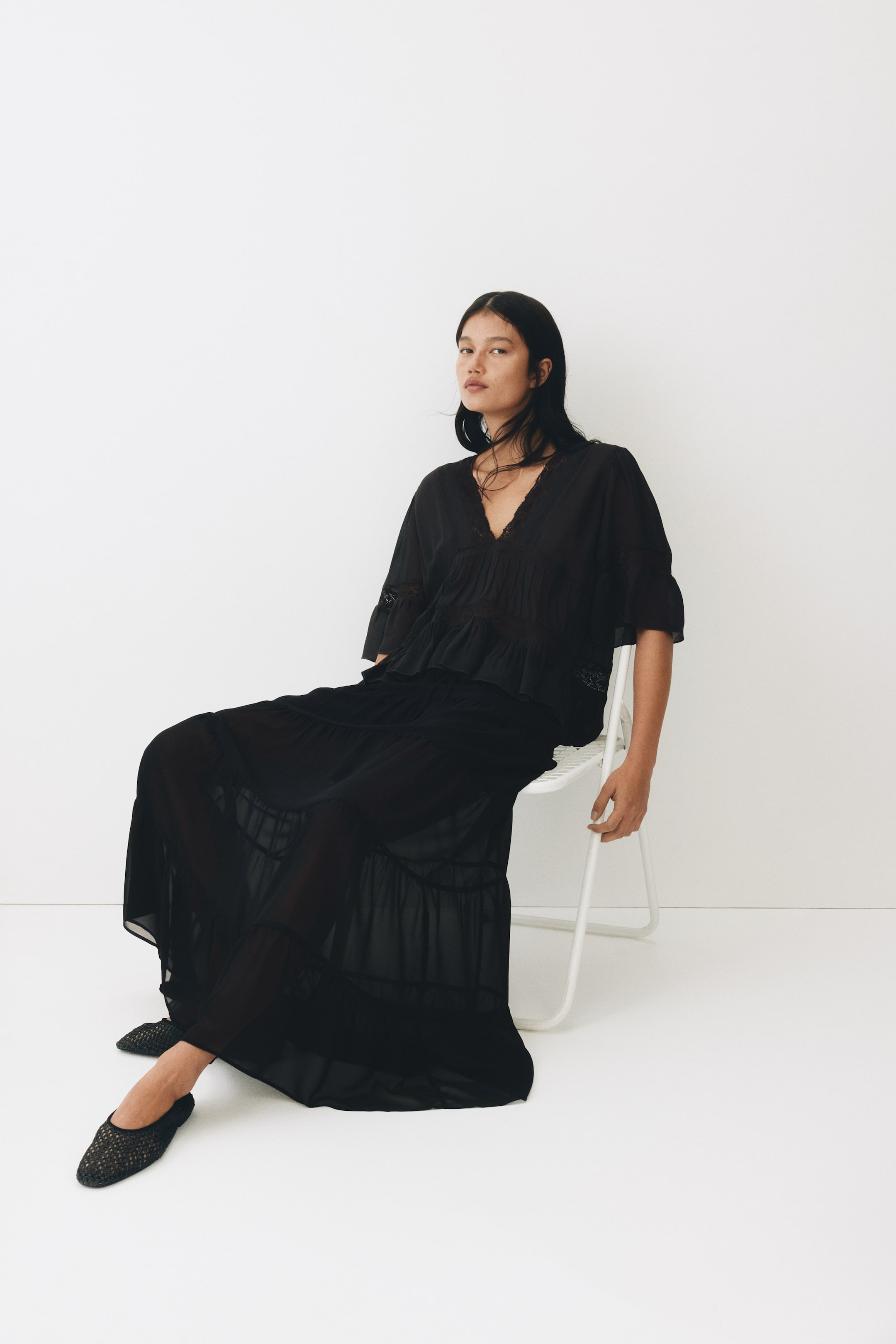 View larger image: A black loose-fit maxi skirt showcases a sheer outer layer and gathered tiers, adding volume. The model is seen wearing the skirt while seated, with the fabric elegantly draping over her legs.