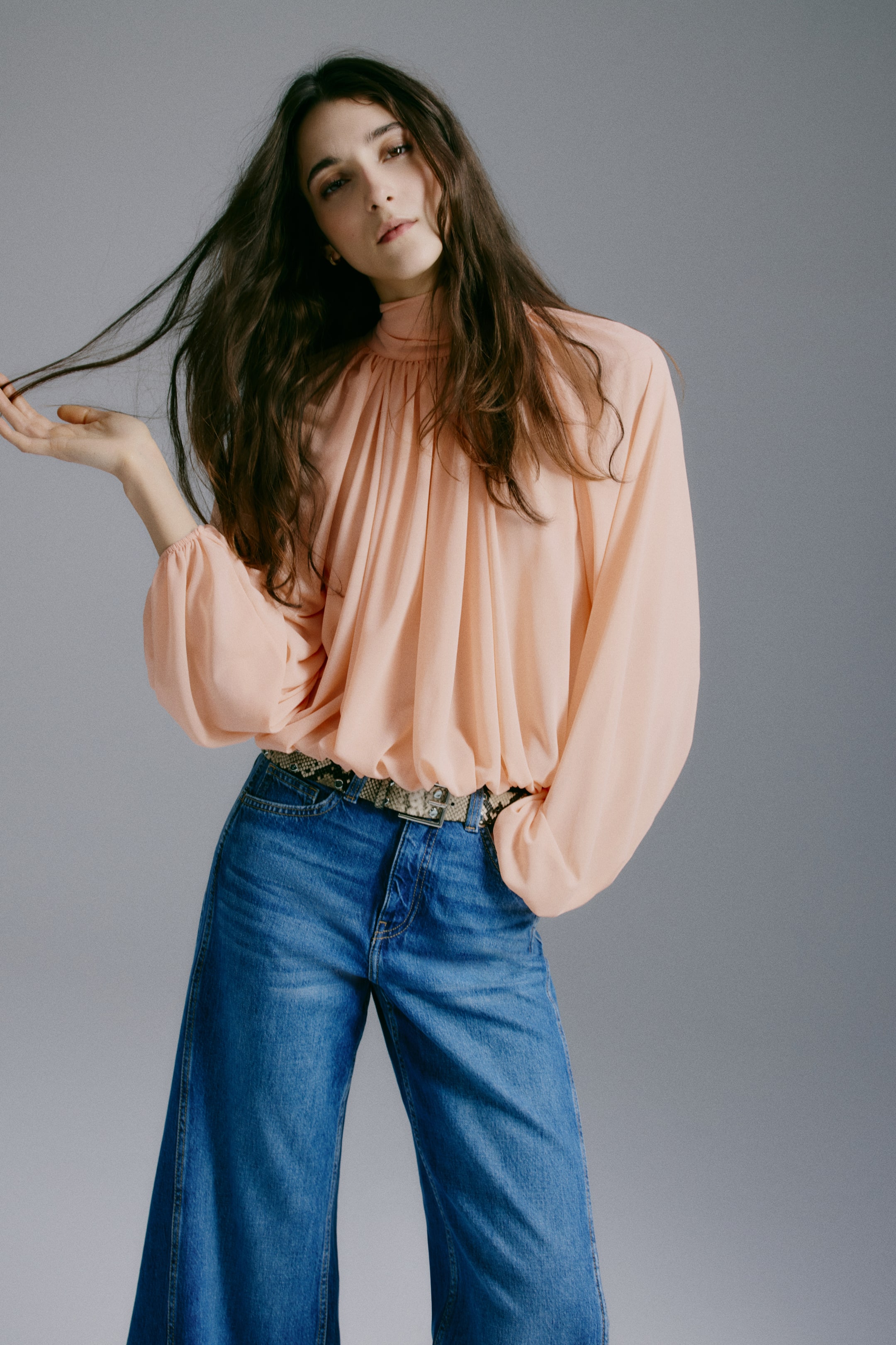 Ampliar la imagen: Una blusa corta y holgada de jersey transparente presenta un cuello alto fruncido y mangas largas abullonadas con puños elásticos. La modelo lleva puesta la blusa, cuyo dobladillo fruncido y elástico crea un efecto drapeado al estar metida en unos jeans azules de pierna ancha y combinada con un cinturón de estampado de serpiente.