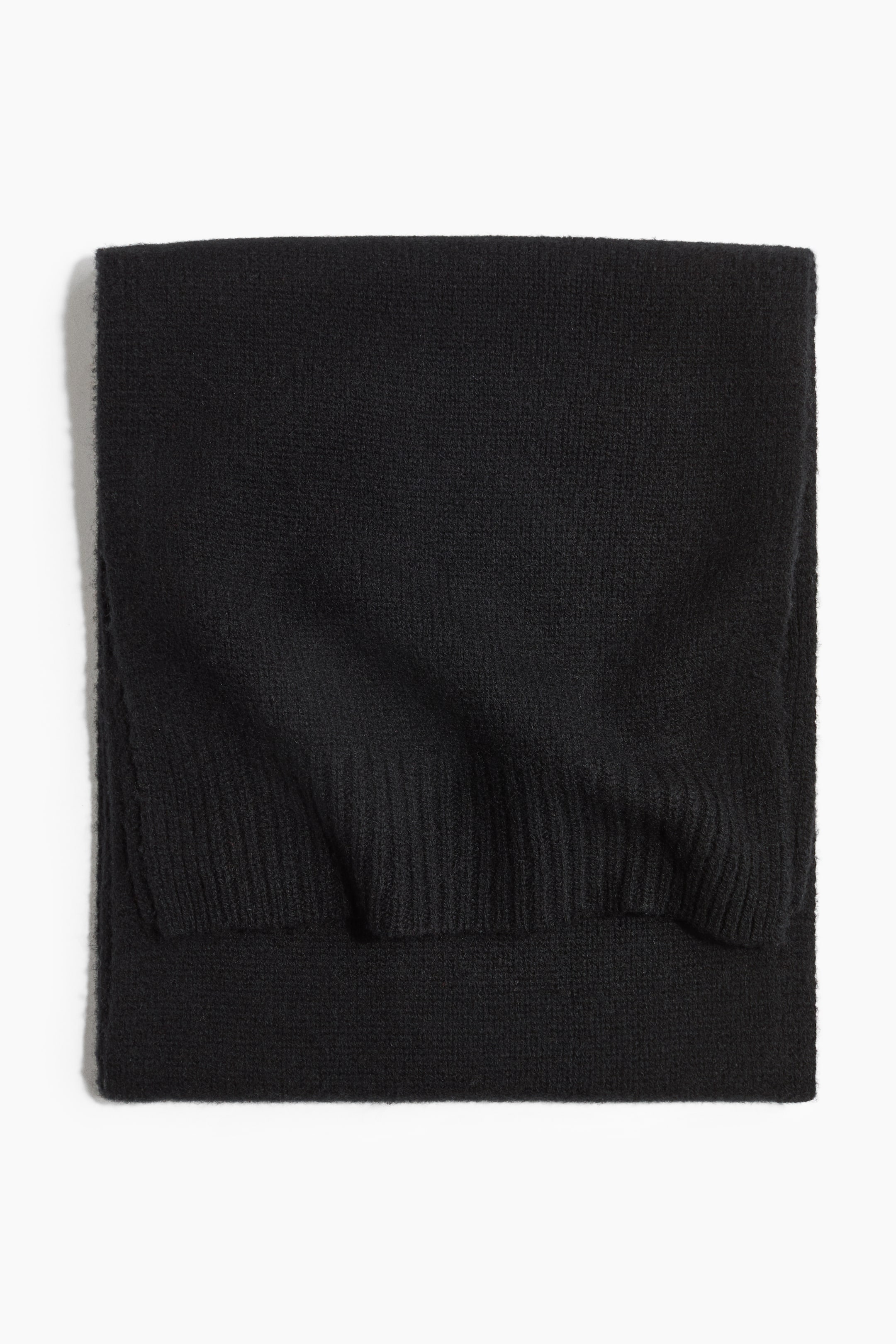Größeres Bild ansehen: Ein schwarzer Strickschal liegt flach gefaltet und zeigt seine weiche, fein gestrickte Struktur sowie einen gerippten Saum an der überlappenden Kante.