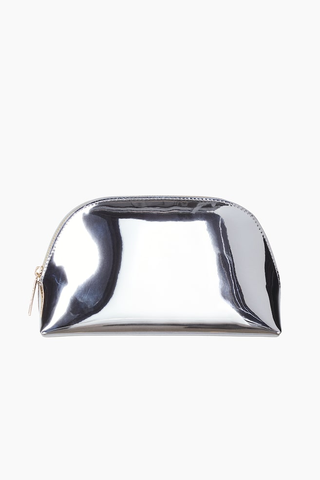 Une trousse de maquillage en forme de dôme est présentée de face, avec une finition argent métallique très réfléchissante et une surface effet miroir. La trousse est conçue avec une base plate et une fermeture zippée incurvée sur le dessus, dotée dâune tirette ton or sur le côté gauche. - 1