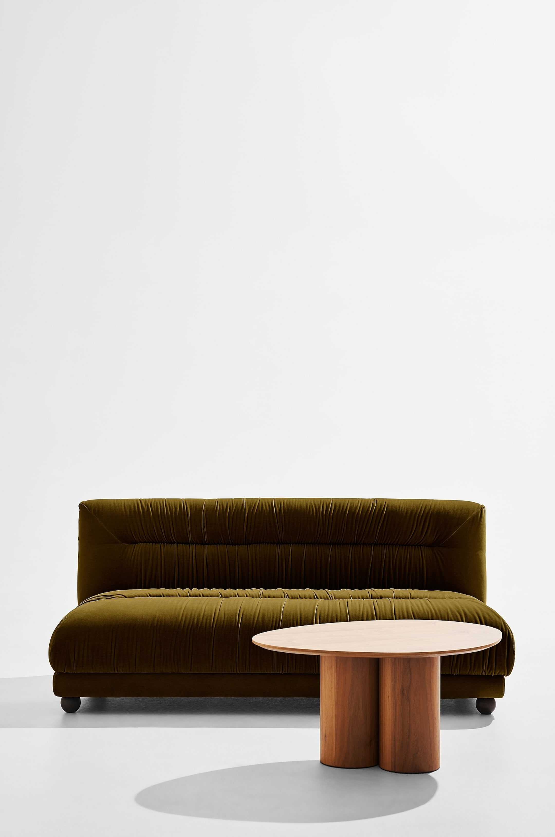 Größeres Bild ansehen: Ein grünes Samtsofa zeigt seine Vorder- und linke Seite und verfügt über ein niedriges Profil mit einer hohen Rückenlehne. Das Sofa präsentiert ein gerafftes Stoffdesign über die Rückenlehne und den Sitz, das horizontale Falten bildet, und ruht auf kleinen, dunklen, runden Füßen.