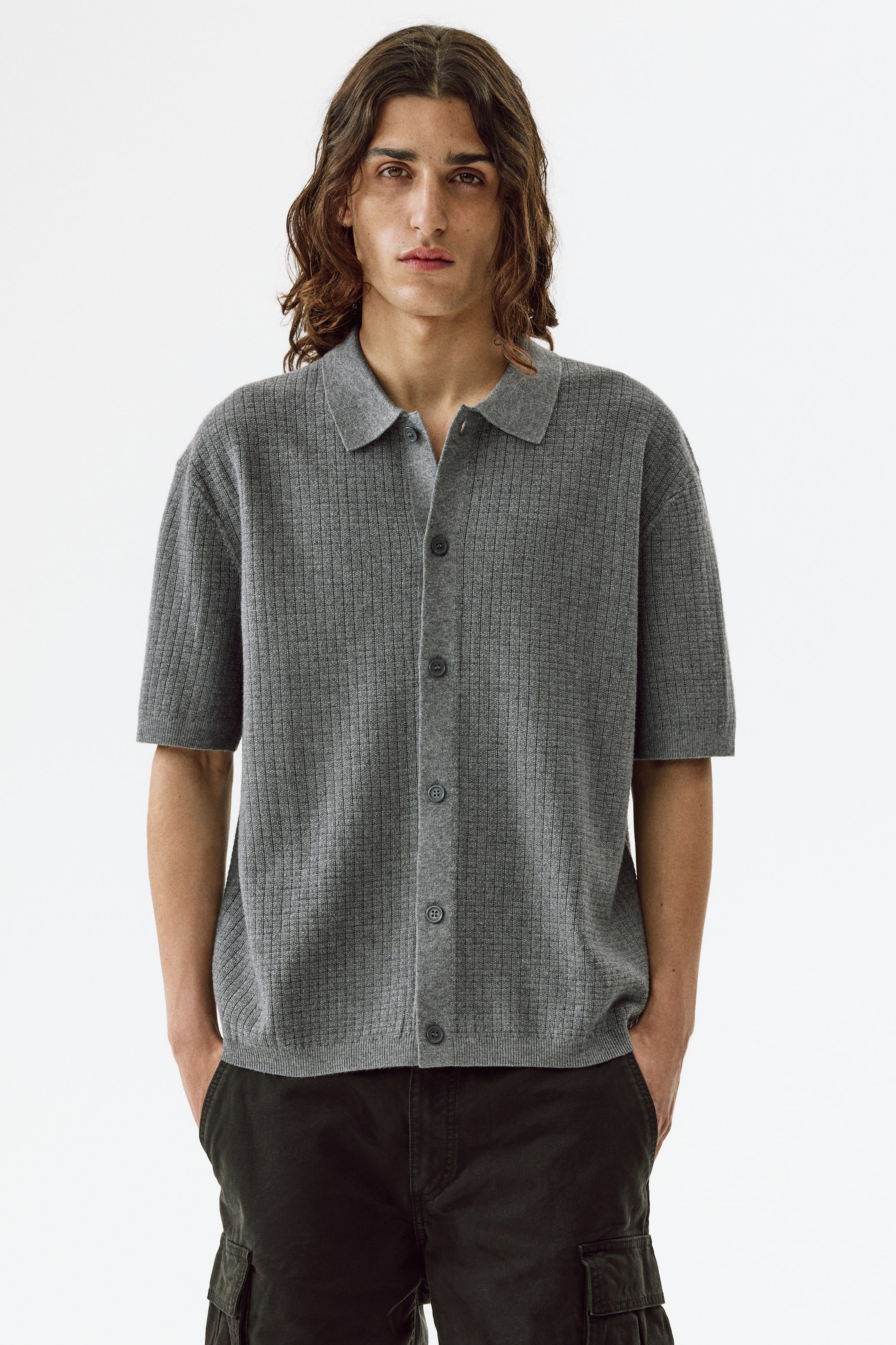 Grotere afbeelding bekijken: Een grijze gebreide poloshirt met een geribbelde textuur, korte mouwen en een klassieke kraag. Het heeft een knoopsluiting aan de voorkant met meerdere donkere knopen.