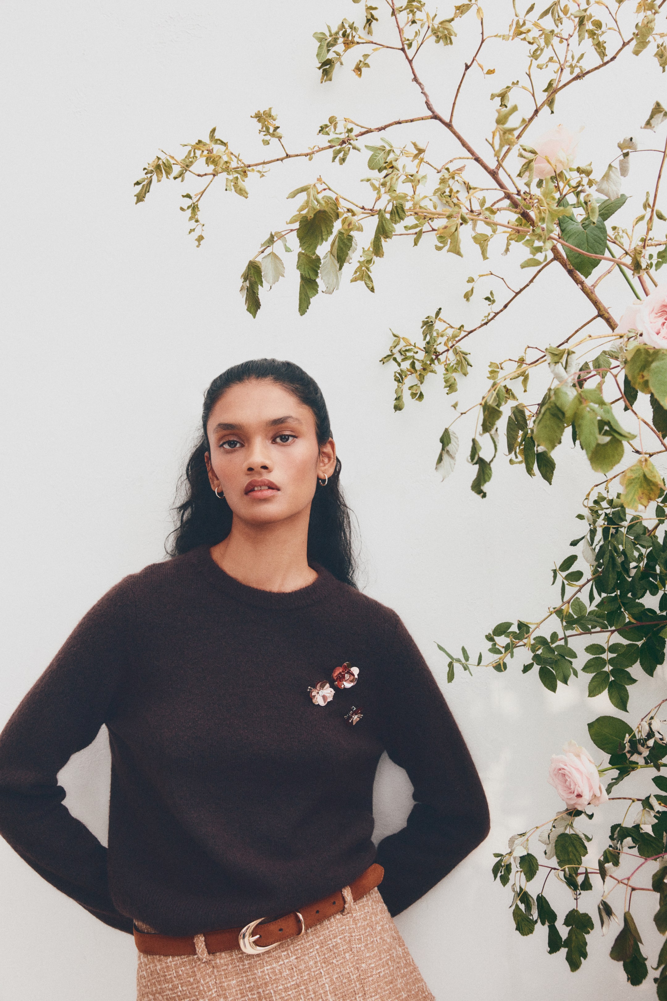 View larger image: A dark brown, textured crew neck sweater is being worn, featuring two small floral brooches on the left chest. It is being styled over a light brown tweed skirt with a brown belt, with the wearer facing slightly to the right.