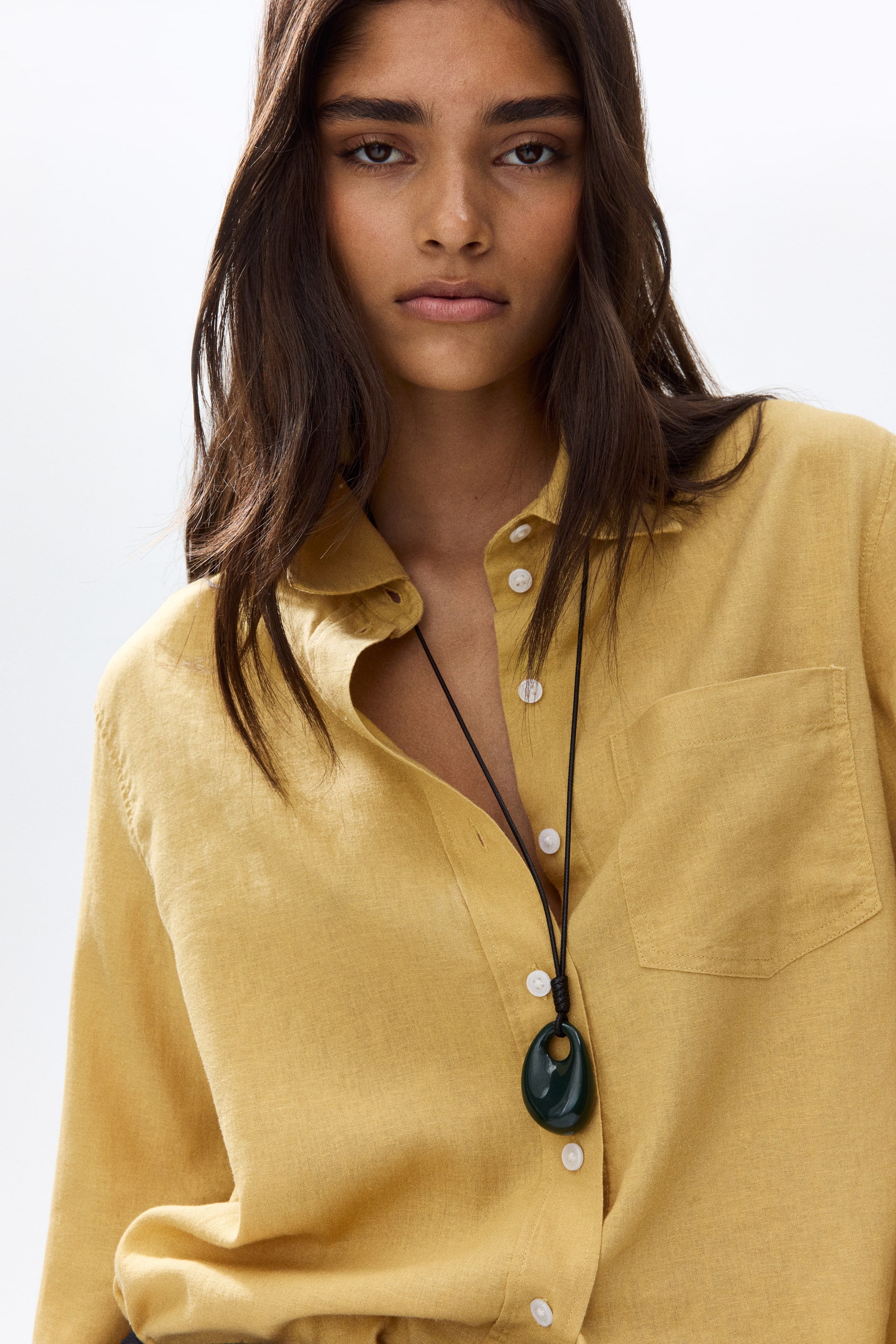 View larger image: The model is donning a relaxed-fit yellow shirt crafted from a breathable linen and viscose blend. It showcases a collar, white buttons running along the front, and an open chest pocket. Complementing the shirt's unbuttoned top is a dark necklace adorned with an oval pendant.