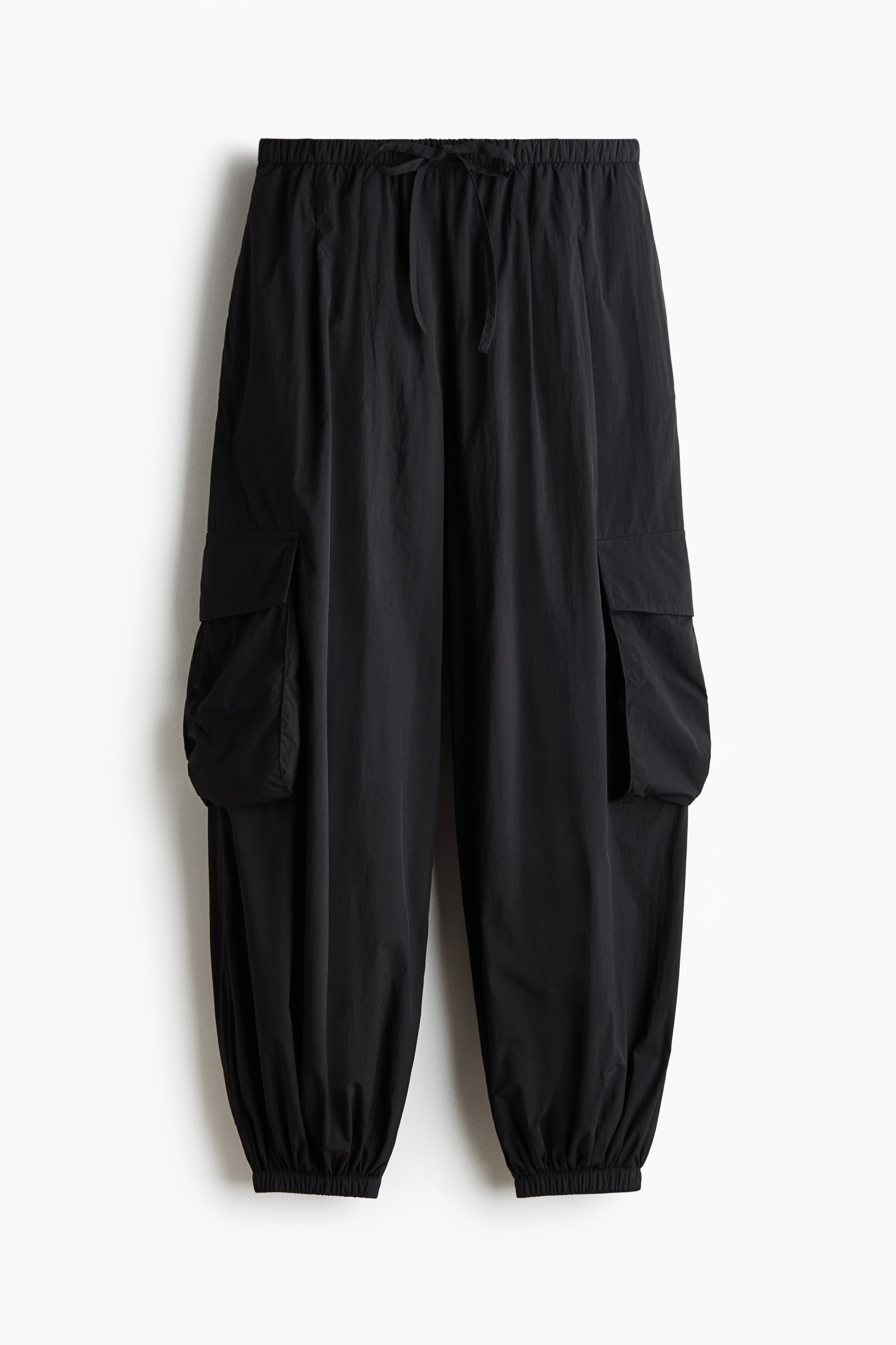 View larger image: The black cargo trousers are showcased in a front view, highlighting an elasticized waistband with a drawstring tie, two spacious cargo pockets with flaps on each leg, and gathered elastic cuffs at the ankles.