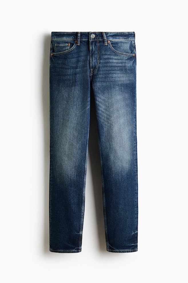 Les jeans en denim bleu sont présentés de face, arborant un délavage bleu moyen avec des décolorations plus claires sur les cuisses. Ils sont conçus avec une taille régulière, une fermeture à bouton, deux poches avant et une poche à monnaie, mettant en valeur une jambe droite et une coupe ajustée de la taille jusqu'à l'ourlet. - 1