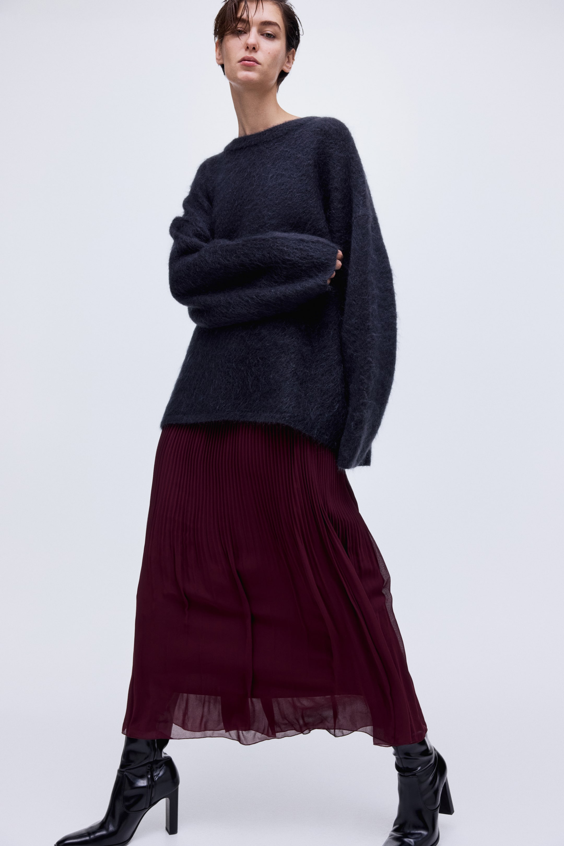 View larger image: A dark grey fuzzy knit crew-neck sweater is worn over a burgundy pleated midi skirt that falls to mid-calf, showcasing a sheer outer layer above black pointed-toe ankle boots.