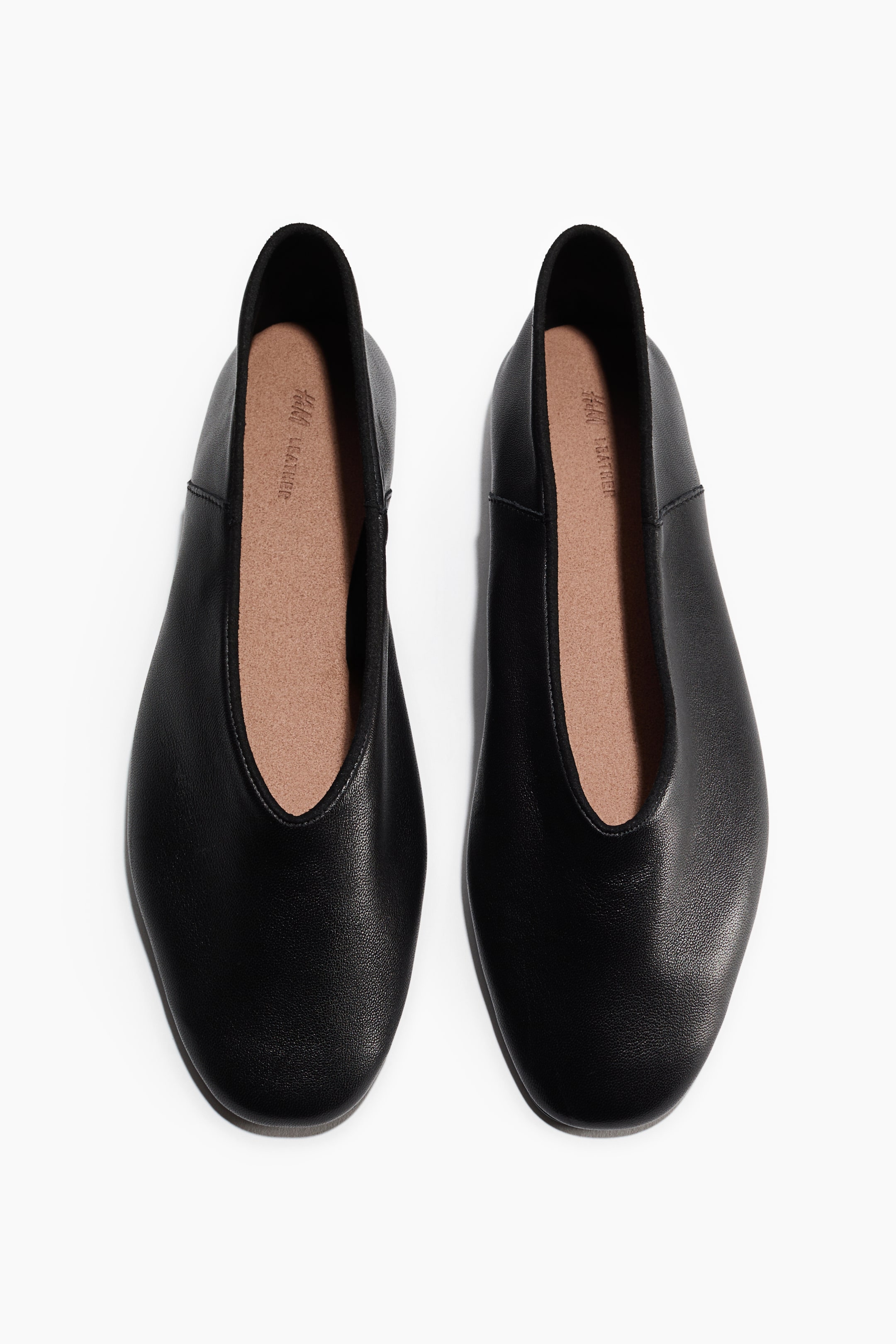 View larger image: Ballet flats crafted from soft black leather with rounded toes are resting side by side in a top-down view, showcasing their deep-cut vamps and textured finish. The flats feature tan insoles and a minimalist slip-on design with subtle side seams.