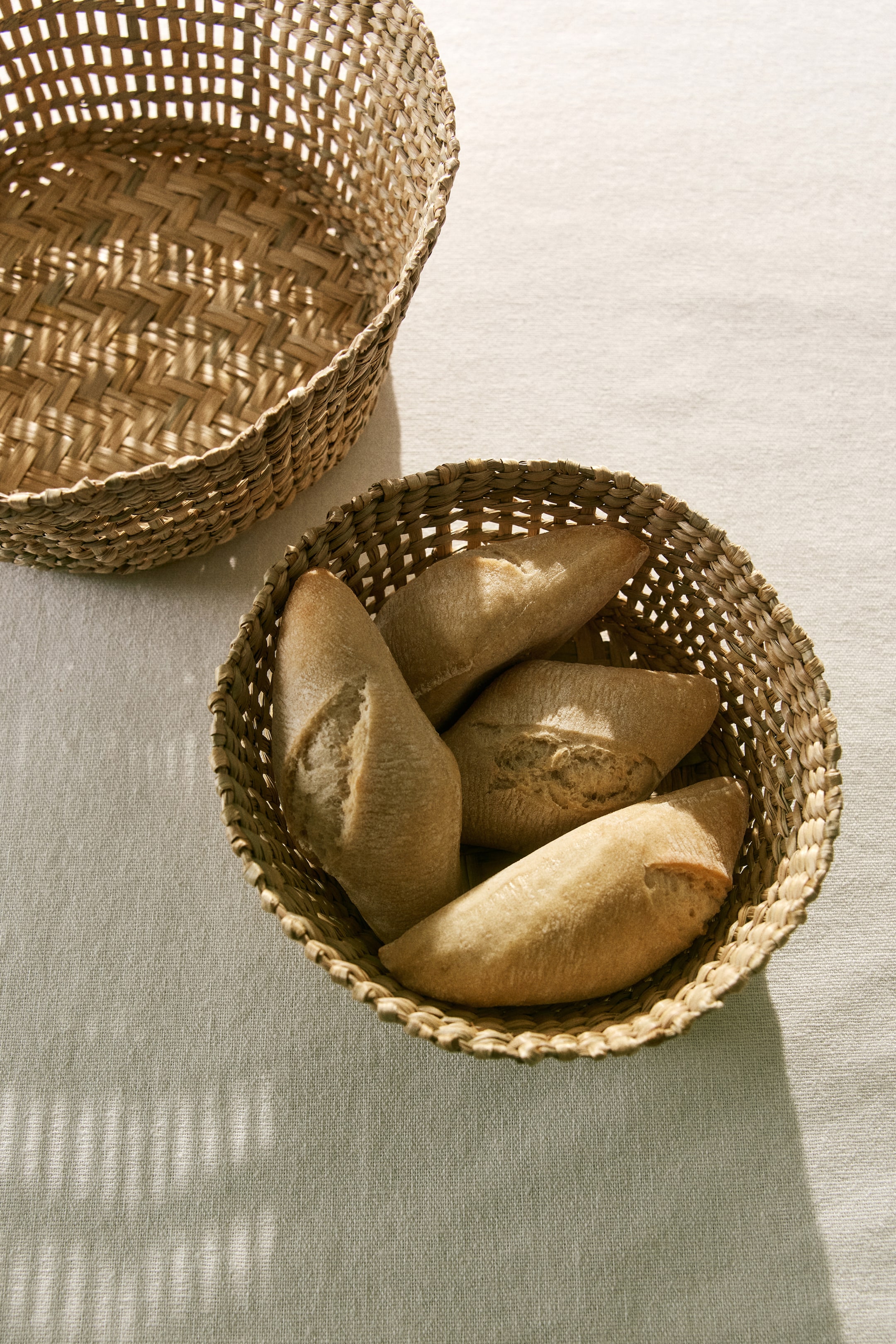 View larger image: "A set of two braided grass baskets is displayed, one larger and one smaller. Both baskets showcase their natural, varied tones and woven texture, with the smaller basket holding several bread rolls."
