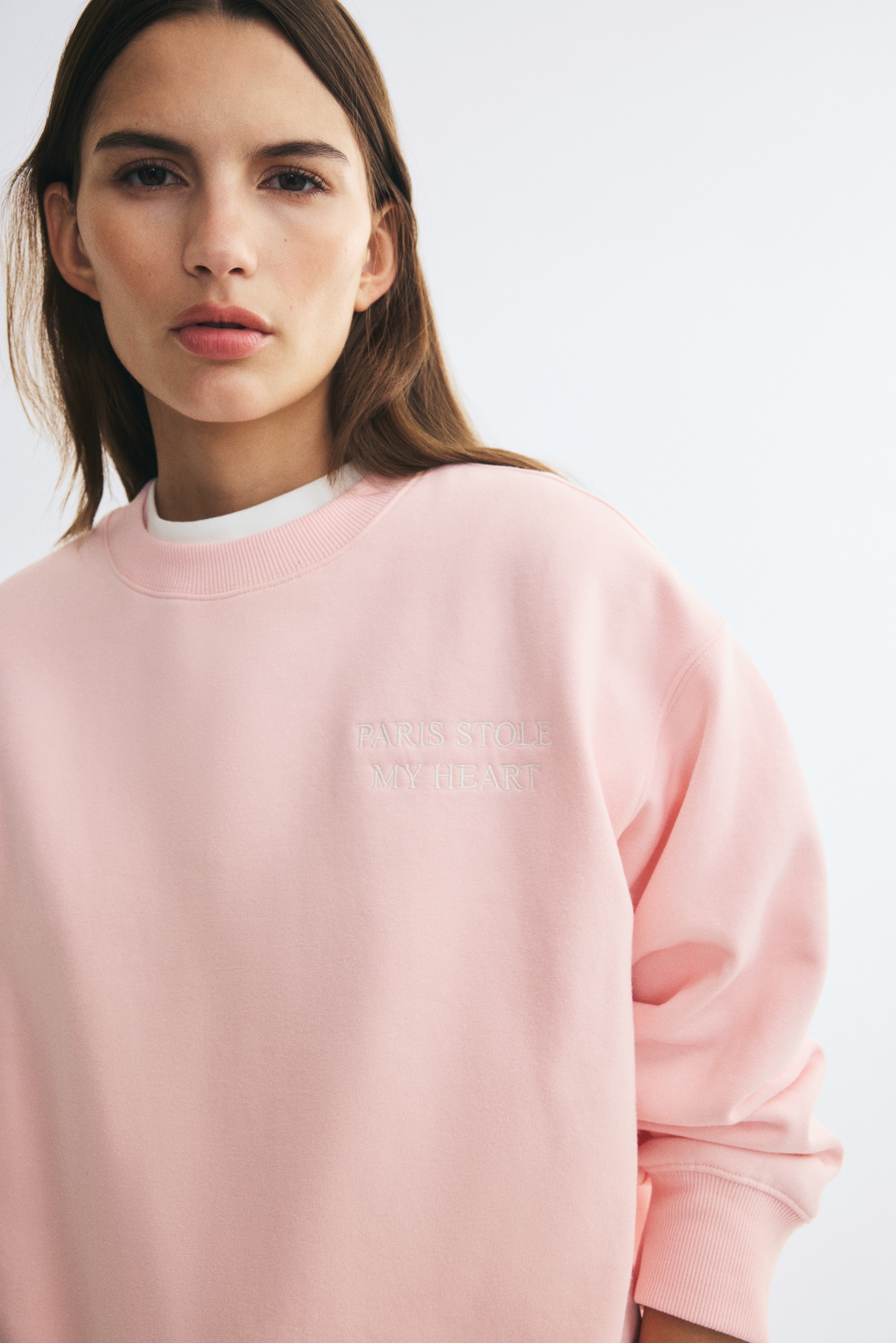 A light pink oversized sweatshirt is hanging in a front view, showcasing a round ribbed neckline, dropped shoulders, and long sleeves with ribbed cuffs. The garment features a ribbed hem and a subtle, tone-on-tone embroidered text design on the upper left chest.