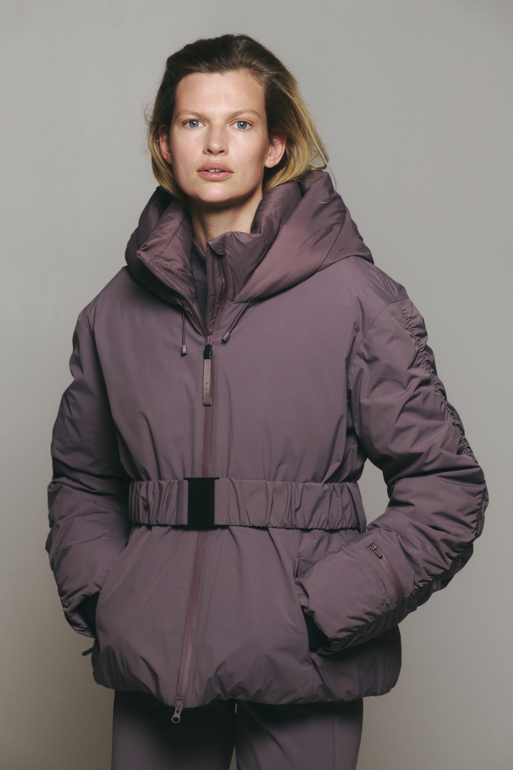 Agrandir l'image: Un mannequin porte une doudoune mauve, les mains glissées dans les poches latérales. La veste présente un col montant, une capuche, une ceinture élastique avec une boucle noire et des détails froncés le long des manches.