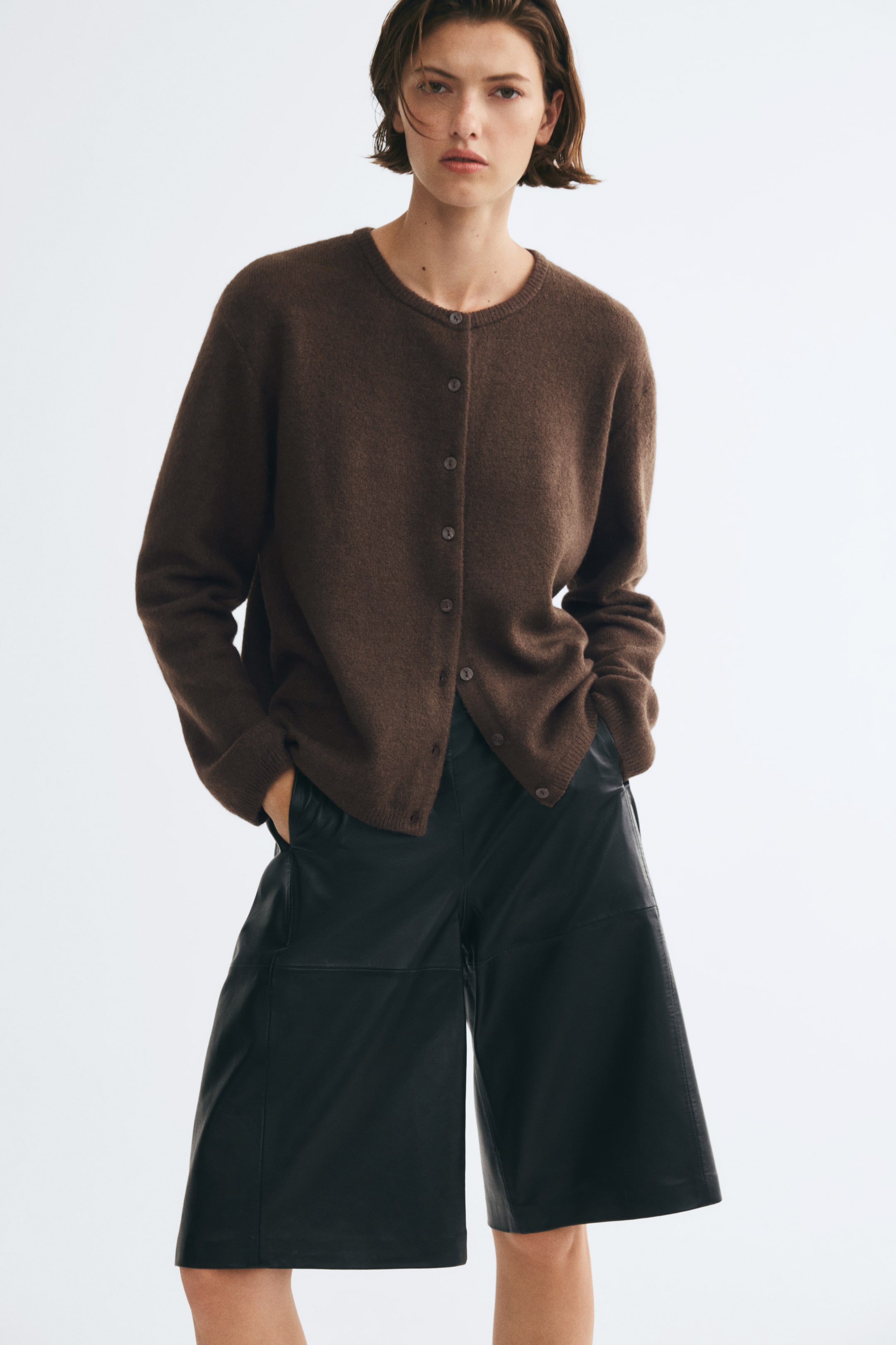 View larger image: A brown, loose-fit knitted cardigan featuring a round neckline, button-through front and dropped shoulders is being worn by the model. The cardigan is styled with wide-leg black leather shorts, with the model’s hands resting in the pockets.